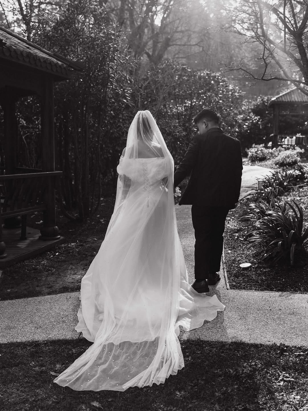 A bride and groom walking together outdoors on a pathway, with the bride in a wedding dress and veil, and the groom in a dark suit, surrounded by trees and bushes, sunlight filtering through the branches.