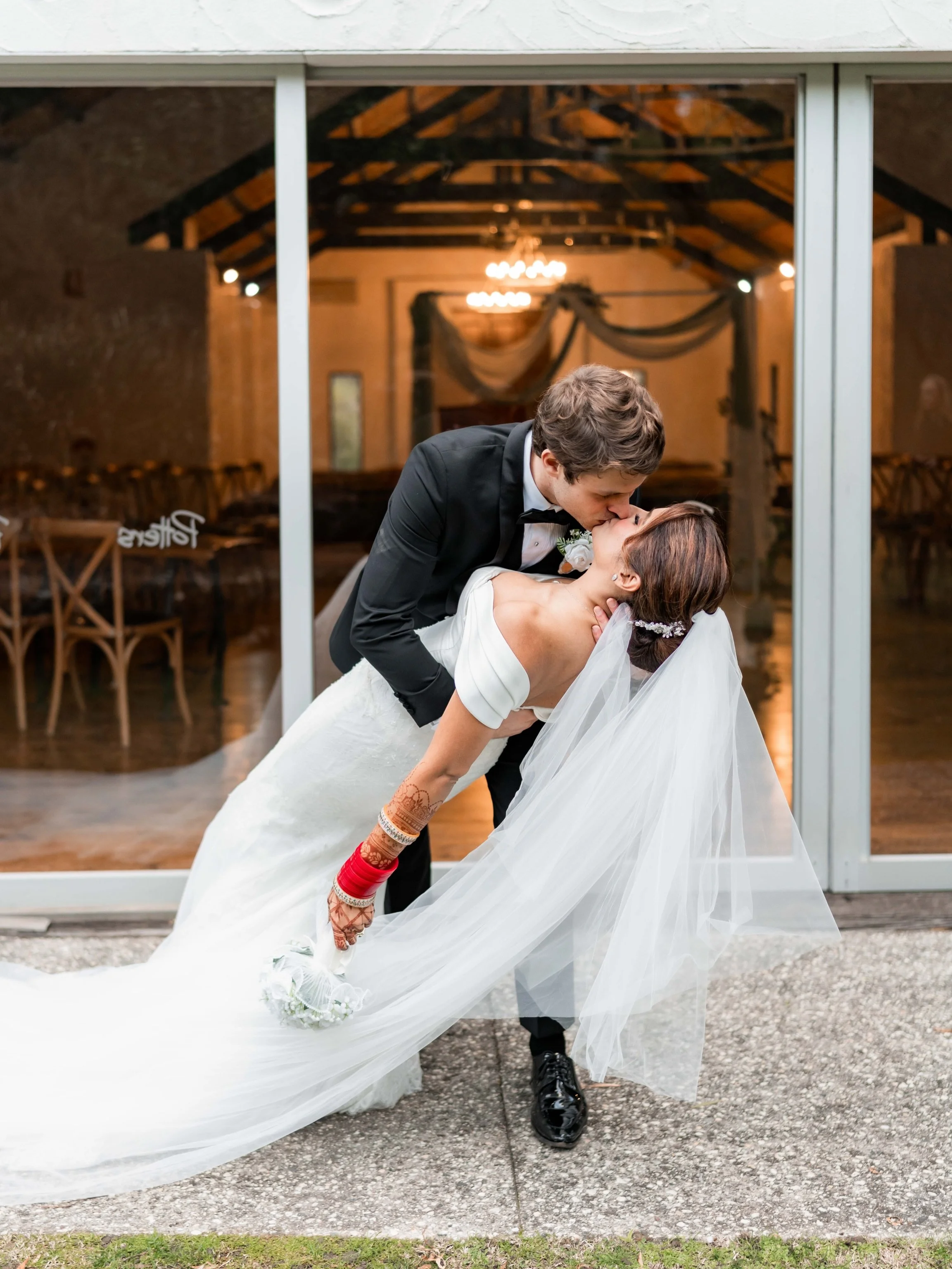 A newlywed couple is kissing through a glass door at a wedding reception venue. The groom is in a tuxedo, bending down to kiss the bride who is in a white wedding dress. The bride's dress is long with a flowing train, and she is wearing traditional h