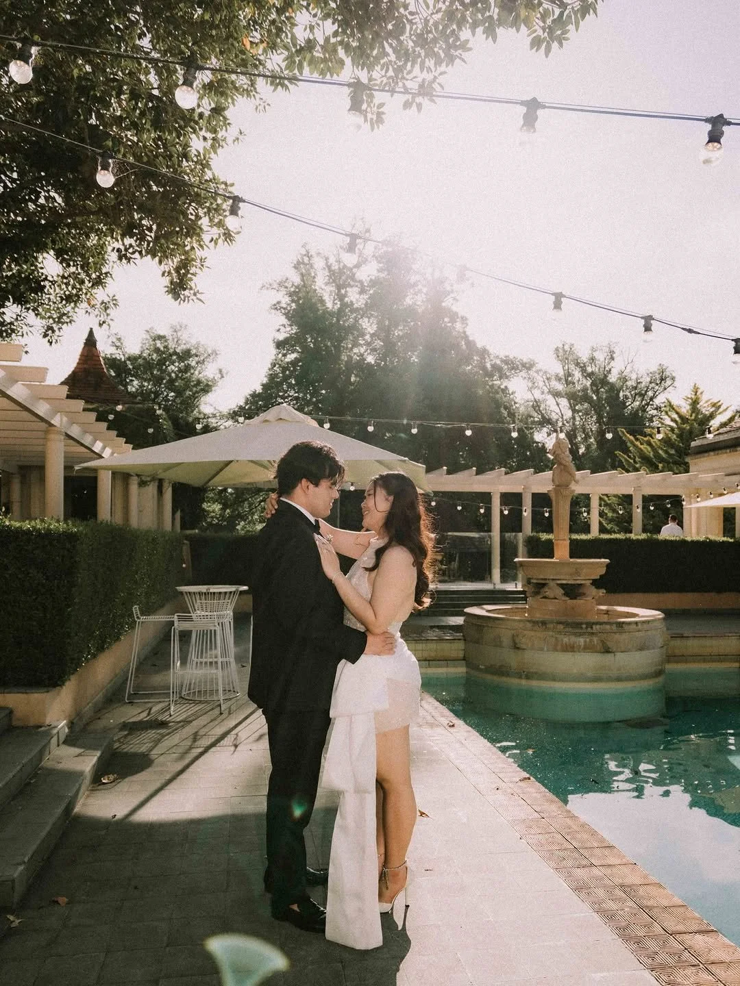A couple in wedding attire dancing by a poolside fountain at sunset, surrounded by trees and outdoor furniture.