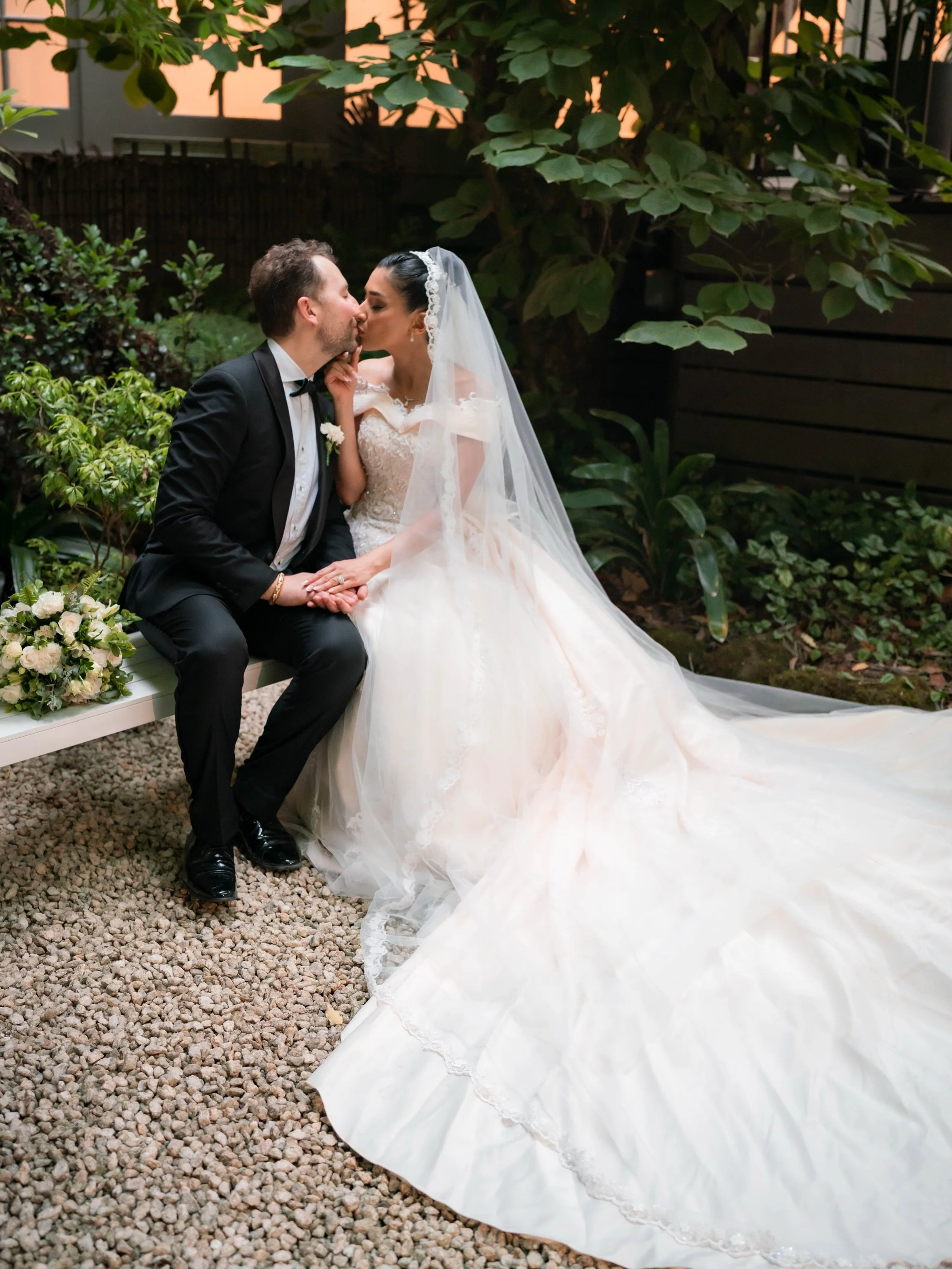 A newlywed couple sharing a kiss, sitting on a white bench in a garden. The groom is wearing a black tuxedo with a bowtie and the bride is wearing a white wedding gown with a veil. Flowers are placed beside them, and a large leafy plant is in the bac