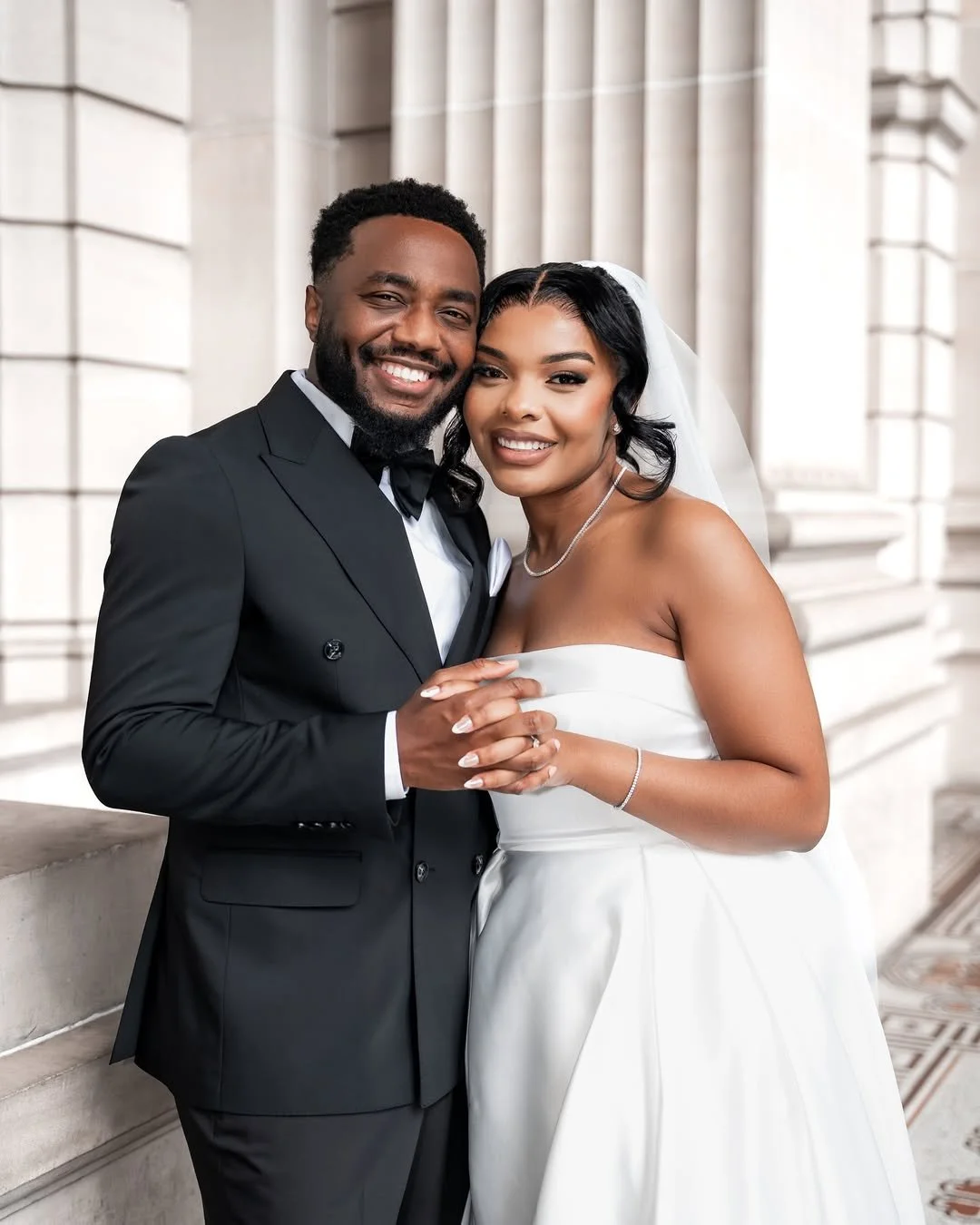 A happy couple on their wedding day, with the groom in a black tuxedo and the bride in a white wedding gown, standing in front of a grand building with columns.