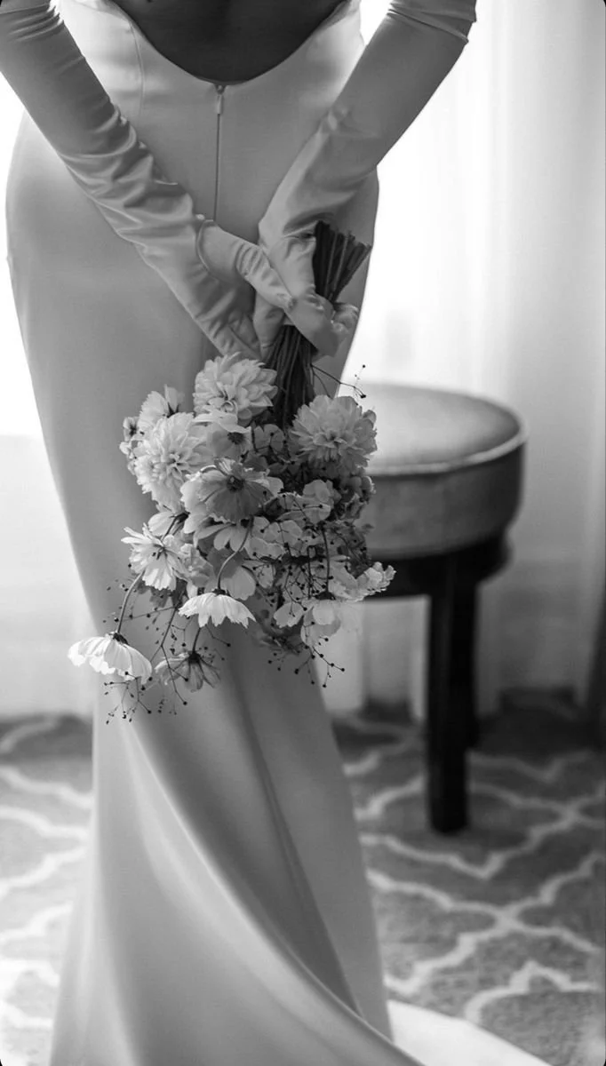 A bride in a long wedding dress holding a bouquet of flowers behind her back, with a small table and patterned carpet in the background.