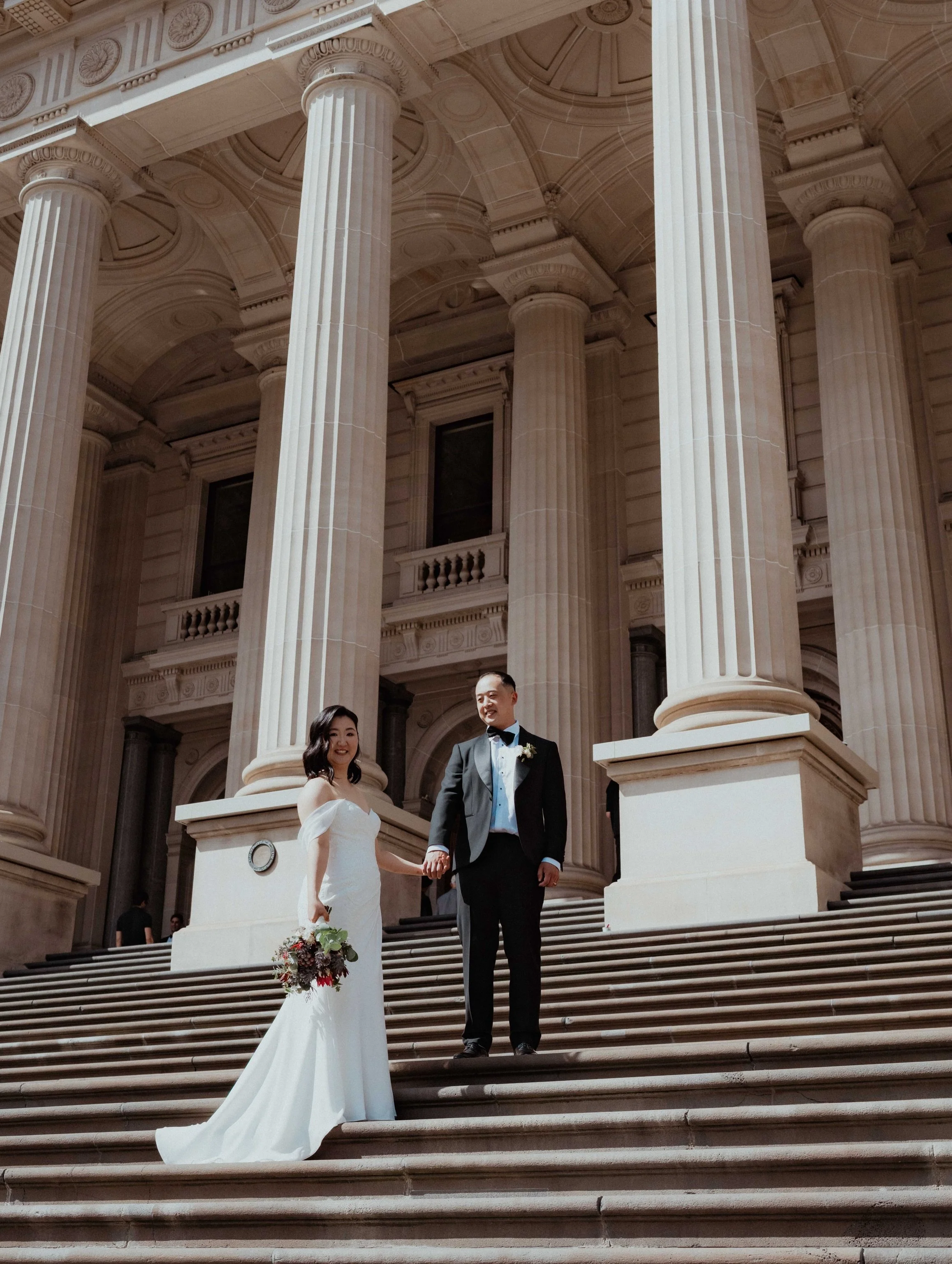 A bride and groom standing on the steps outside a grand building with tall columns, holding hands and smiling, during a wedding ceremony.