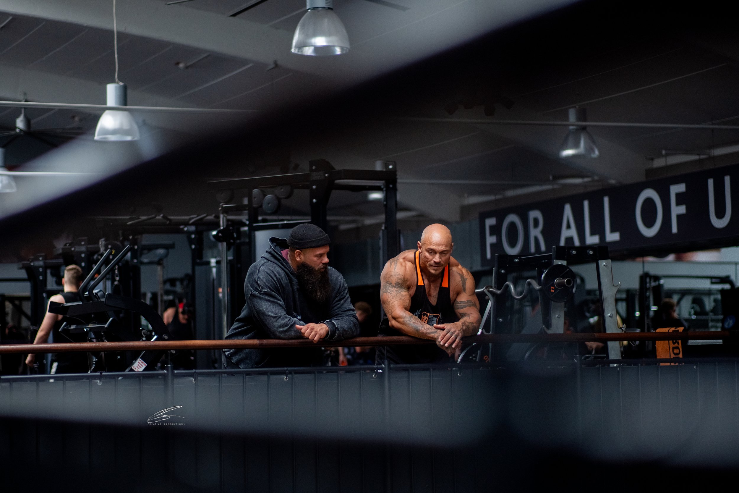 Zwei Männer in einem Fitnessstudio im Gespräch, einer trägt einen schwarzen Tanktop und Shorts, der andere trägt eine schwarze Kapuzenjacke und eine Mütze.