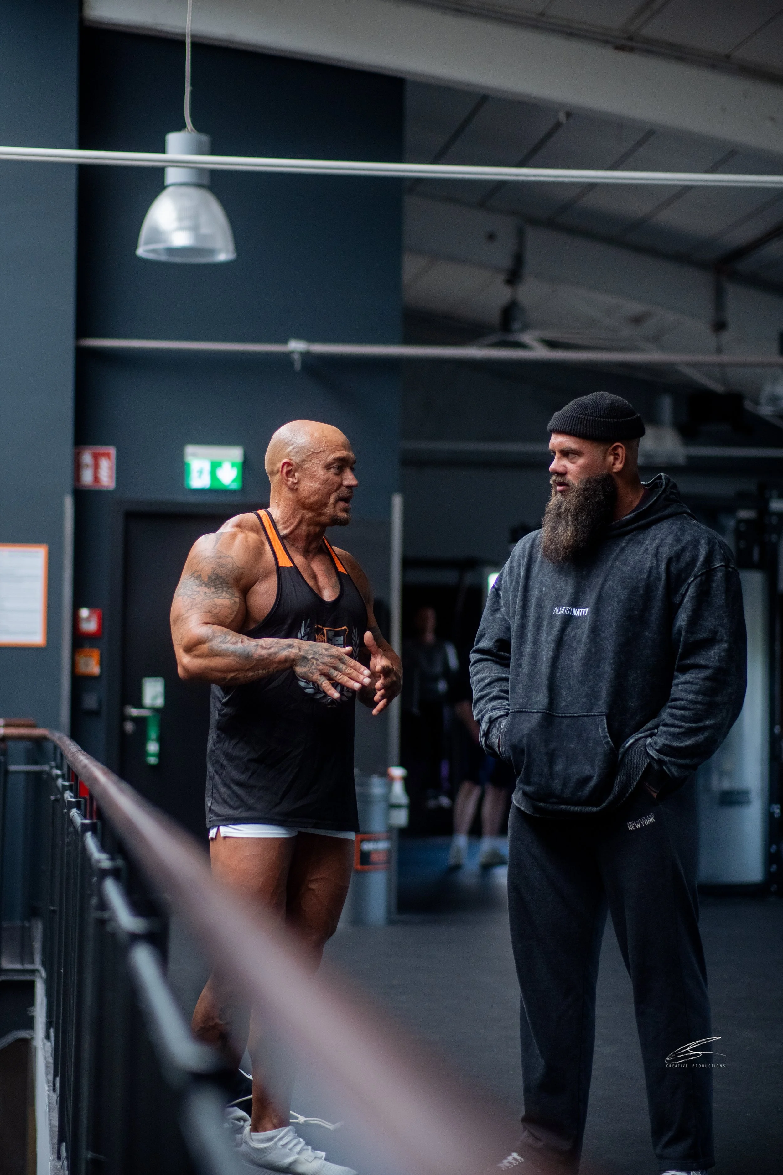 Zwei Männer in einem Fitnessstudio im Gespräch, einer trägt einen schwarzen Tanktop und Shorts, der andere trägt eine schwarze Kapuzenjacke und eine Mütze.