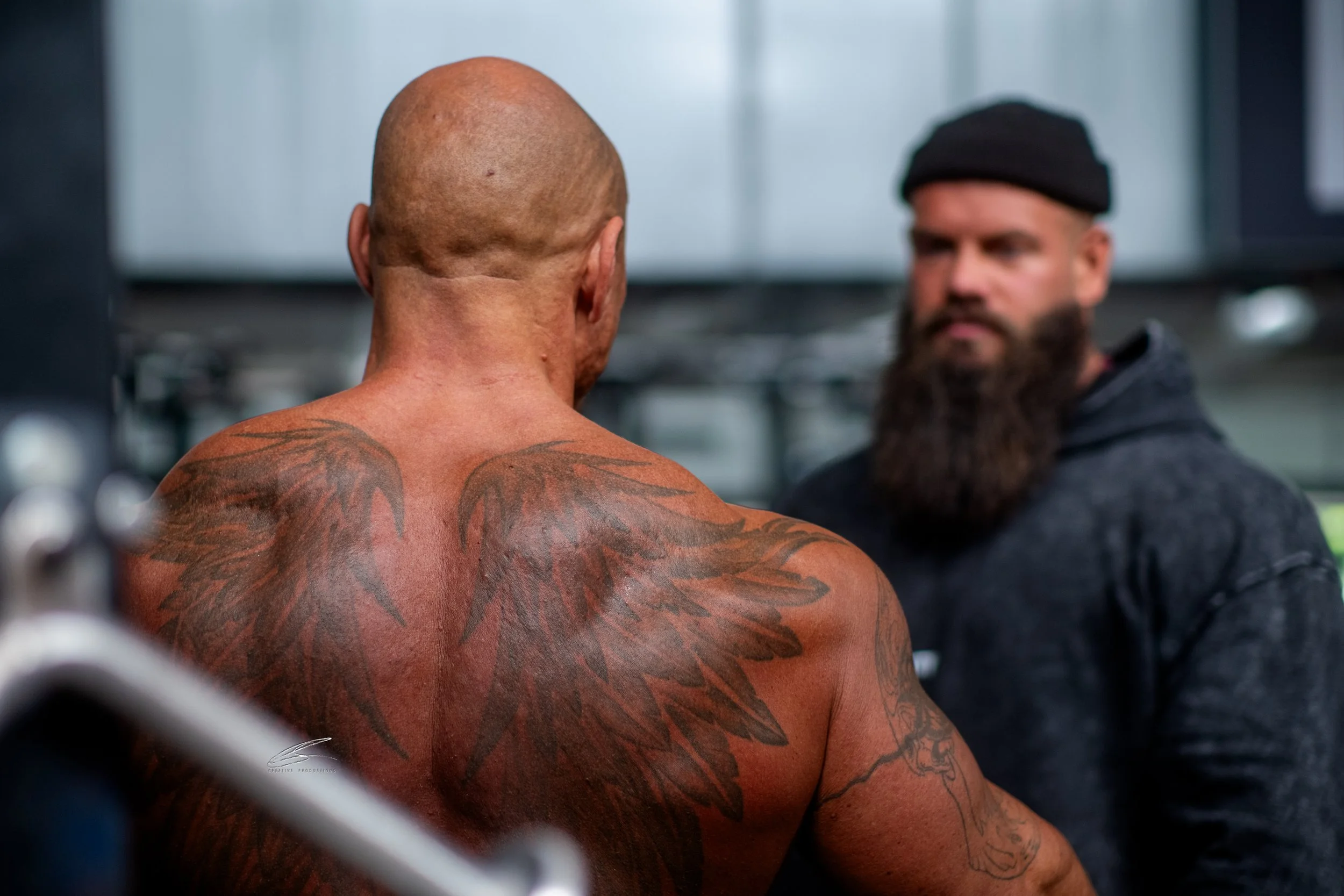 Ein muskulöser, tätowierter Mann mit glattem Kopf sitzt in einem Fitnessstudio, während eine andere Person mit einem Bart und einer schwarzen Mütze ihm gegenübersitzt.