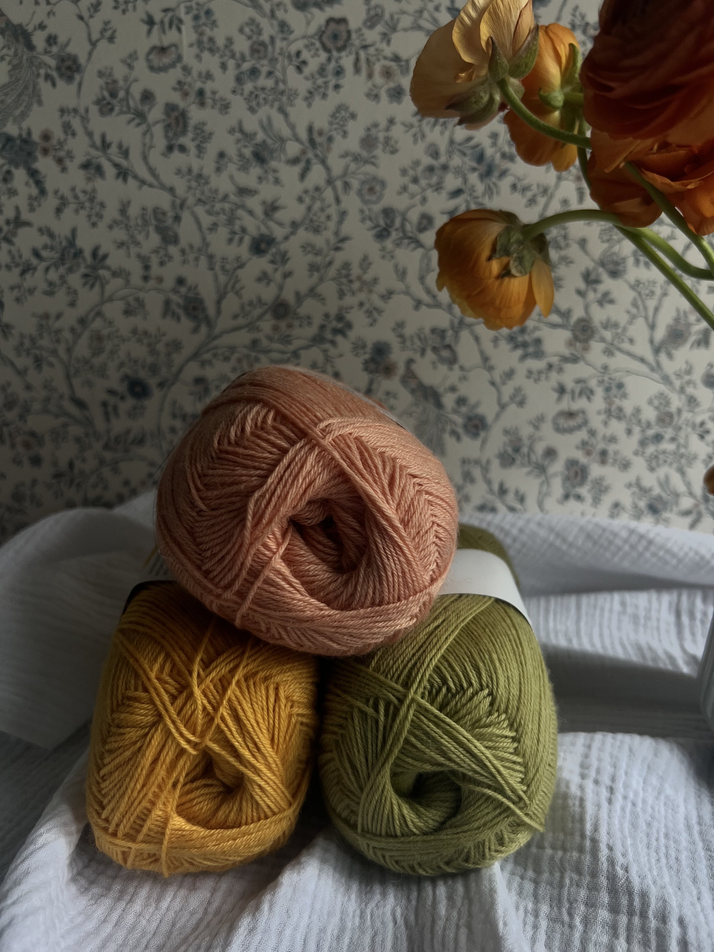 Drei Knäuel Garn in den Farben Rosa, Gelb und Grün stehen auf einem weißen Tuch vor einer blumigen Tapete. Im Hintergrund sind gelbe Blumen.