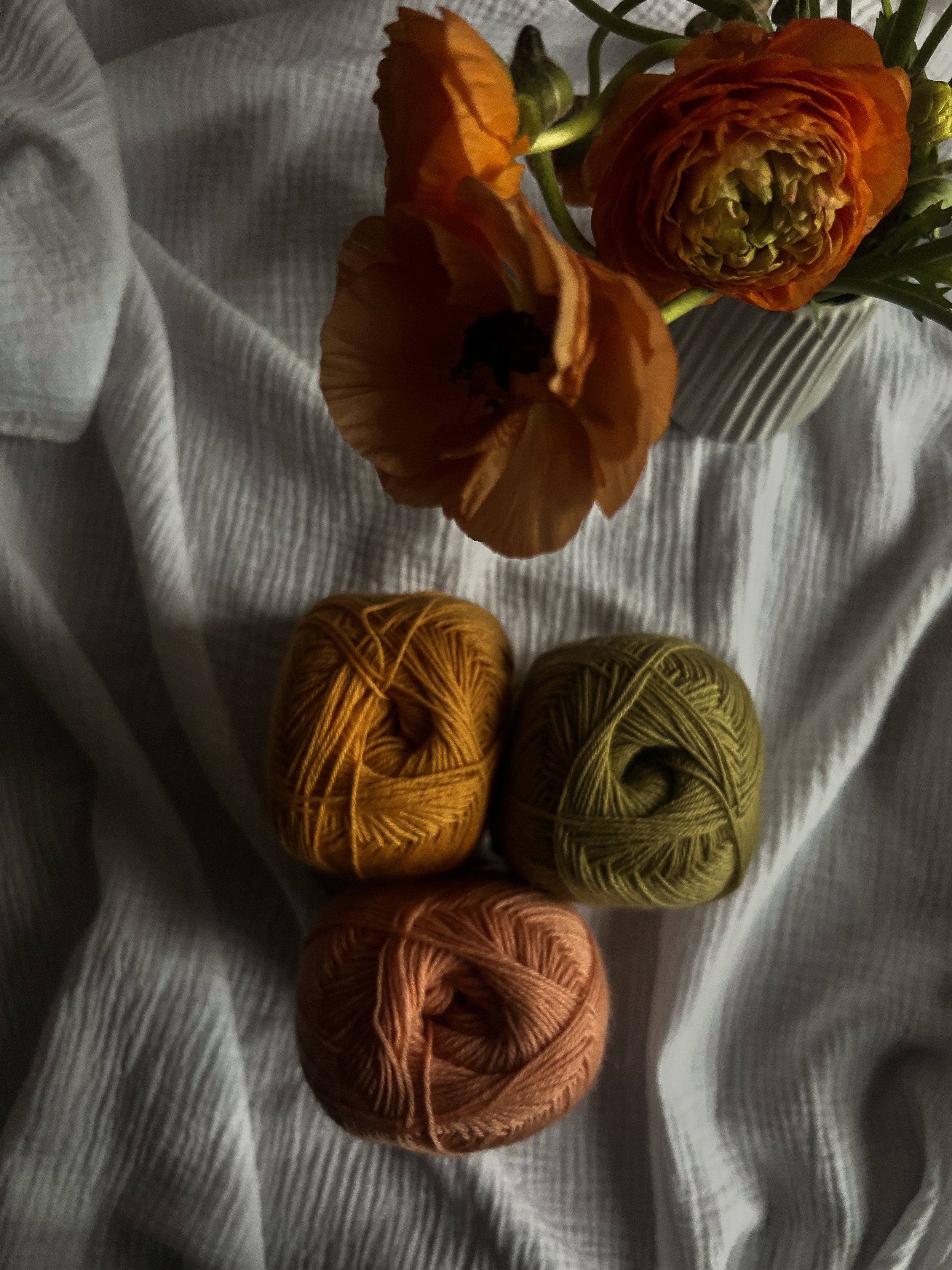 Blick auf eine Vase mit orangefarbenen Blumen und drei Knäuel Wolle in den Farben Orange, Grün und Rosa, die auf einem weißen Stoff liegen.