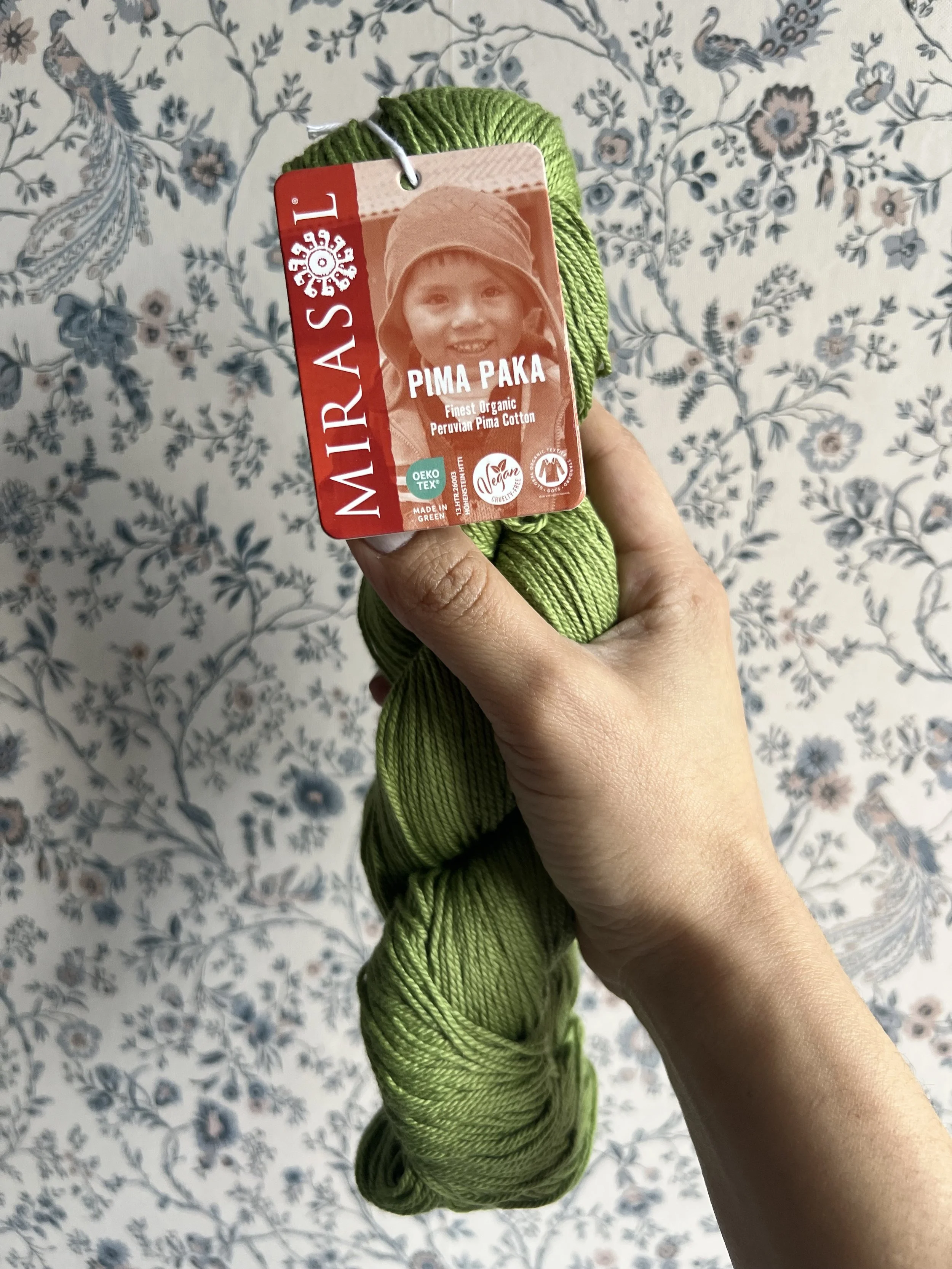 Hand hält grünes Knäuel Wolle mit Etikett, Hintergrund mit blauen und weißen Blumenmuster.