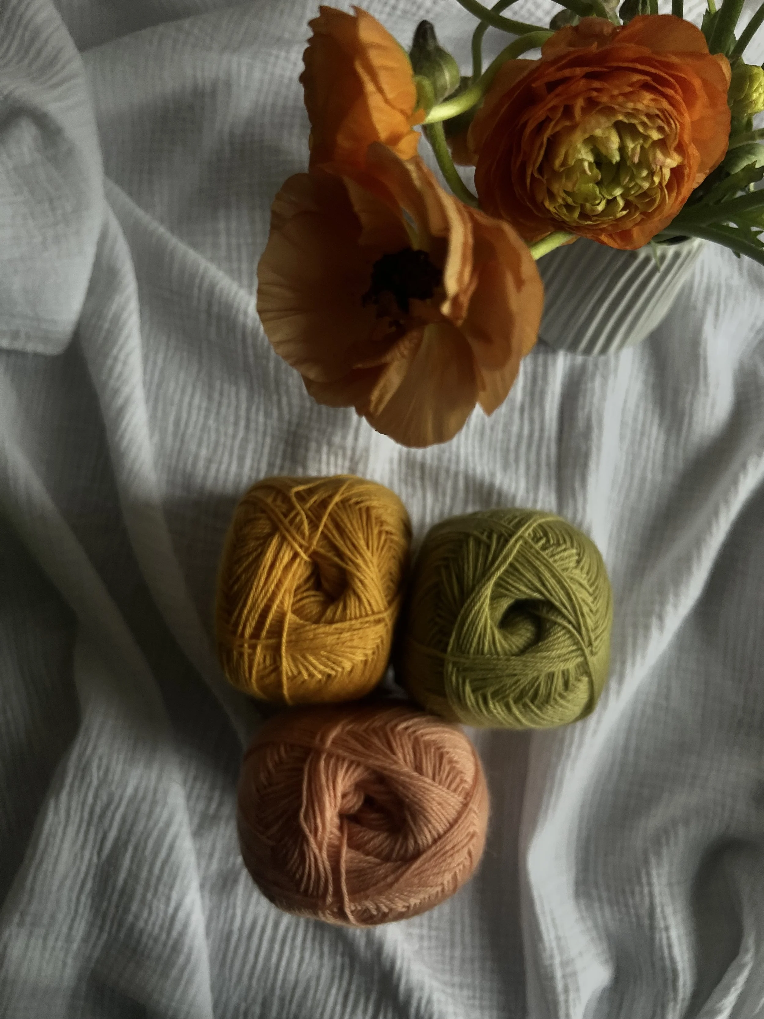 Bunte Blumen in einer Vase neben drei gegenstände, die Garnknäuel in gelb, grün und pfirsichfarben, auf einem weißen Stoff.