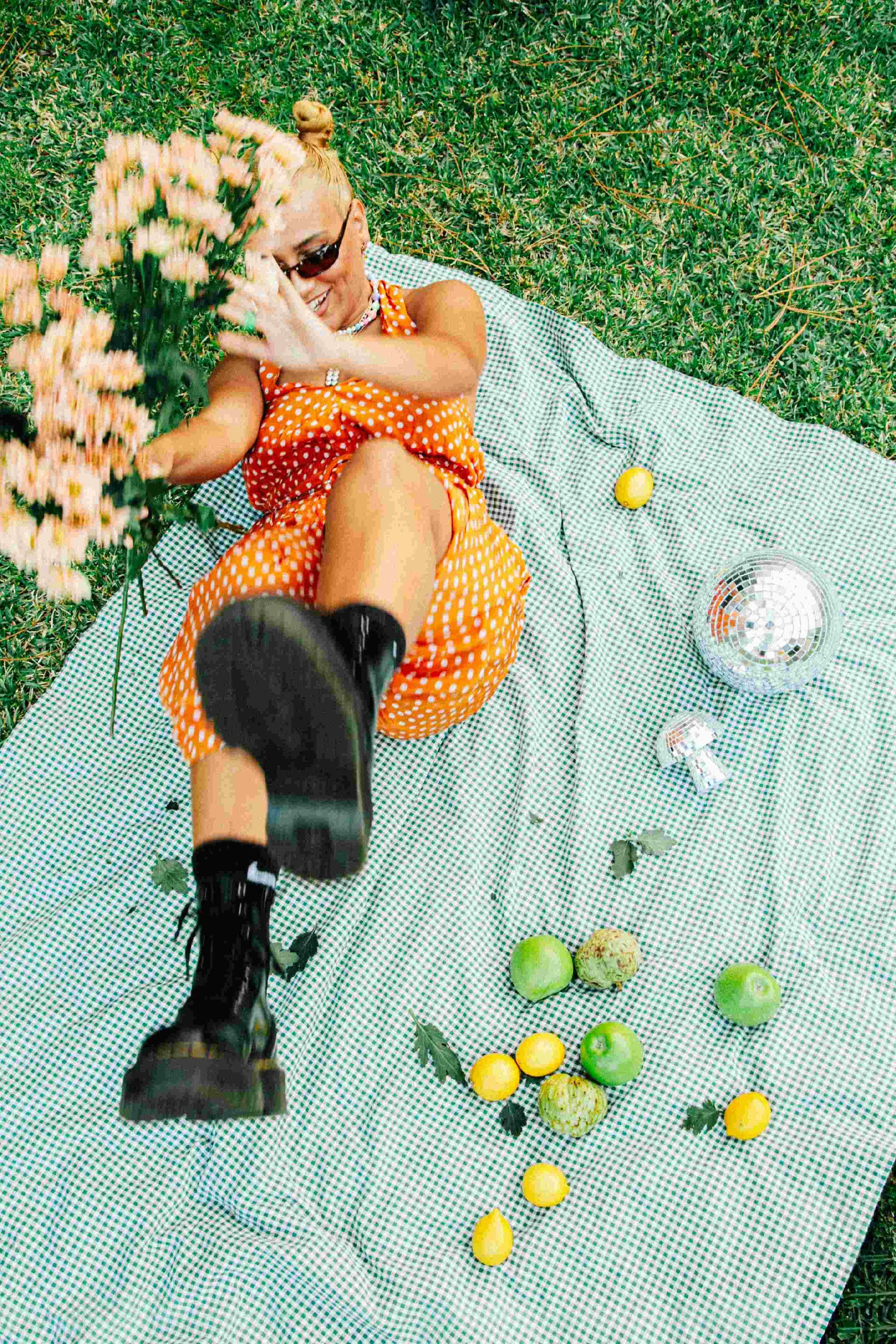 Mixed blonde woman with space buns, lying on a picnic blanket in a grassy area, wearing sunglasses, Dr. Martens boots and an orange polka dot dress, laughing and throwing flowers, with green apples, yellow lemons, a disco ball.