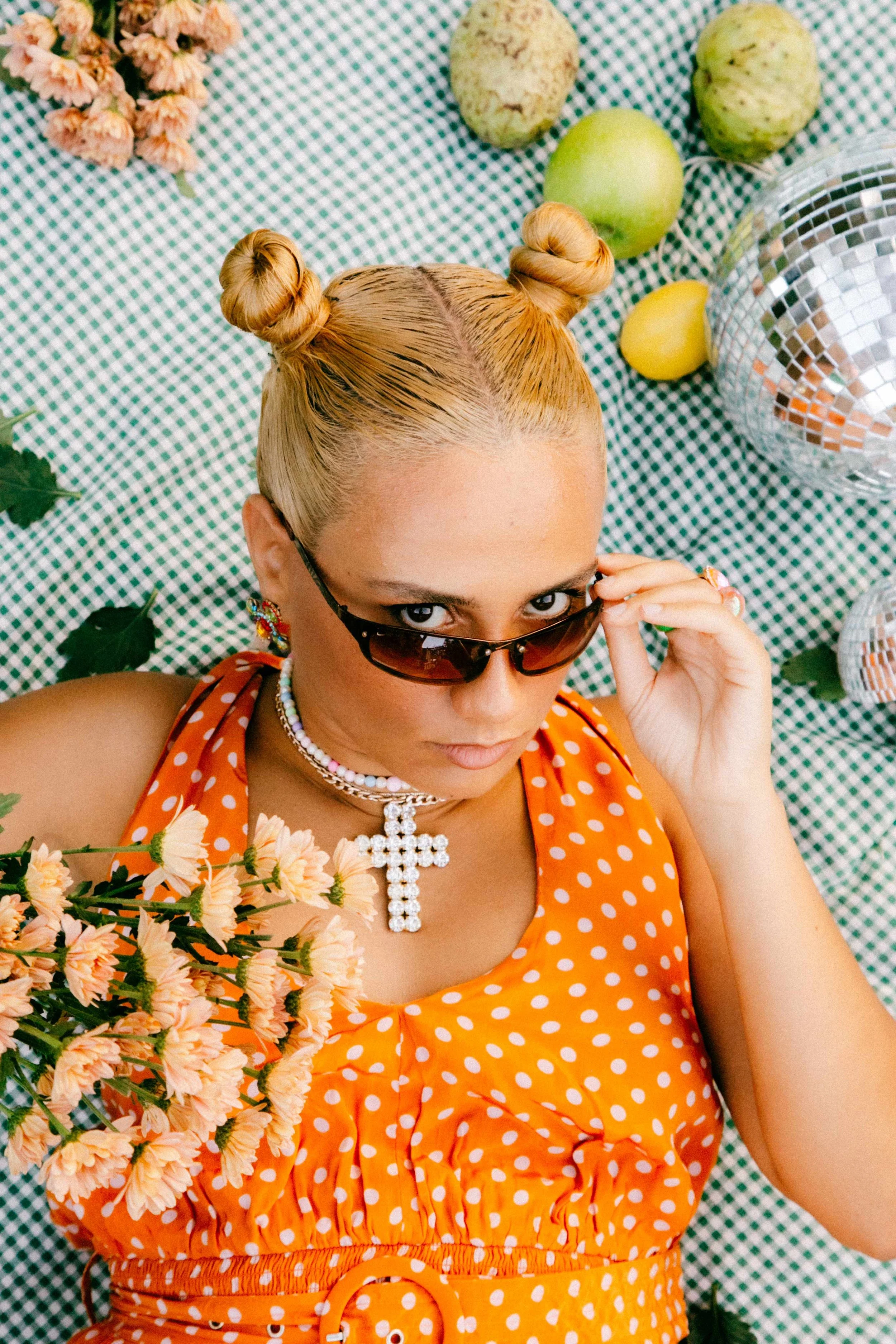 Mixed blonde woman with space buns, wearing sunglasses, with an orange polka dot dress, and a cross necklace, lying on a checkered green and white tablecloth surrounded by light orange flowers, yellow lemons, green apples, and disco balls.