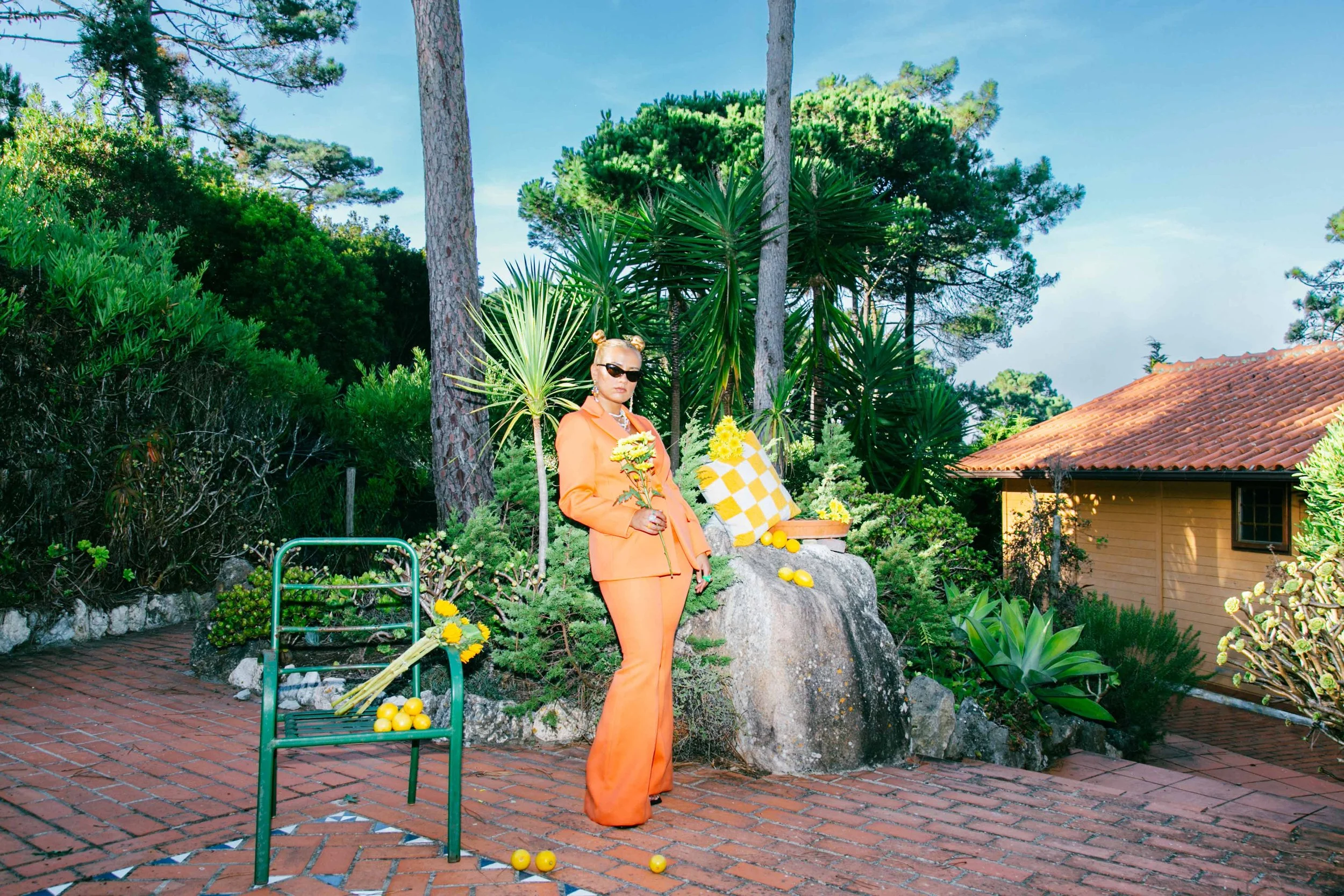 Mixed black African and white American stylish and cool blonde woman with space buns, dressed in an orange suit and sunglasses standing outdoors with a summer yellow lemon-themed decor, surrounded by lush green plants and tall trees under a blue sky.