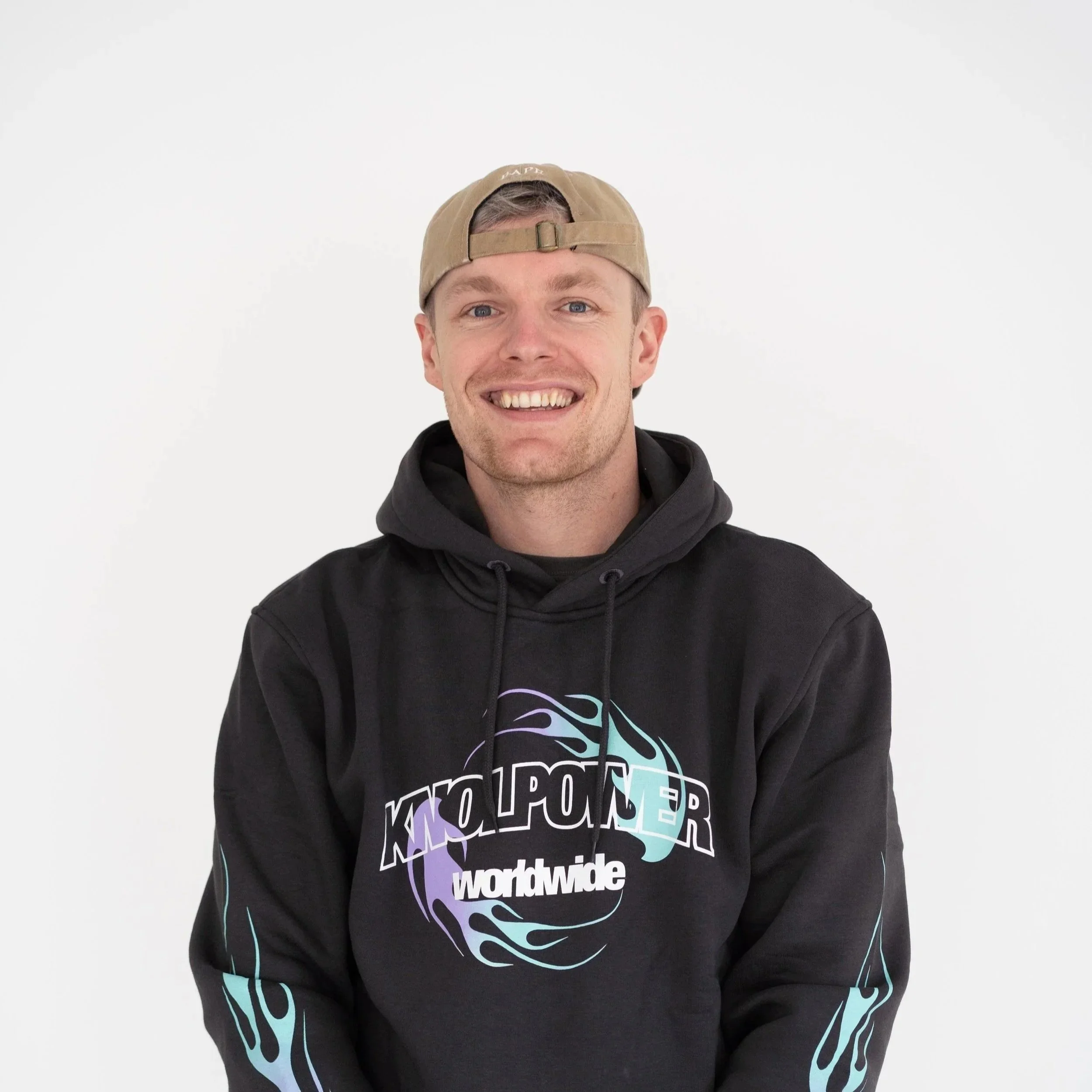 Smiling man wearing a beige backward cap and a black hoodie with colorful graphic design and text that reads 'worldwide'
