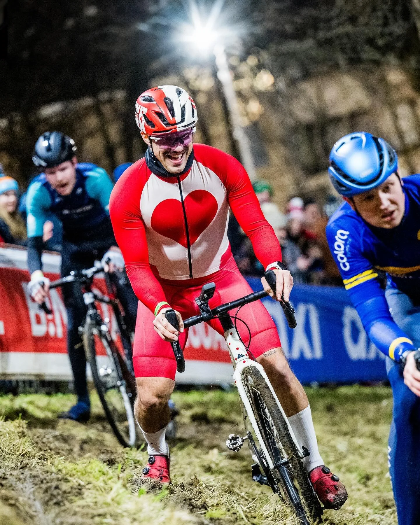 Cyclist in a red and white jersey with a heart design, wearing a red helmet and pink glasses, smiling while riding a mountain bike on a dirt trail at night with other cyclists in the background.