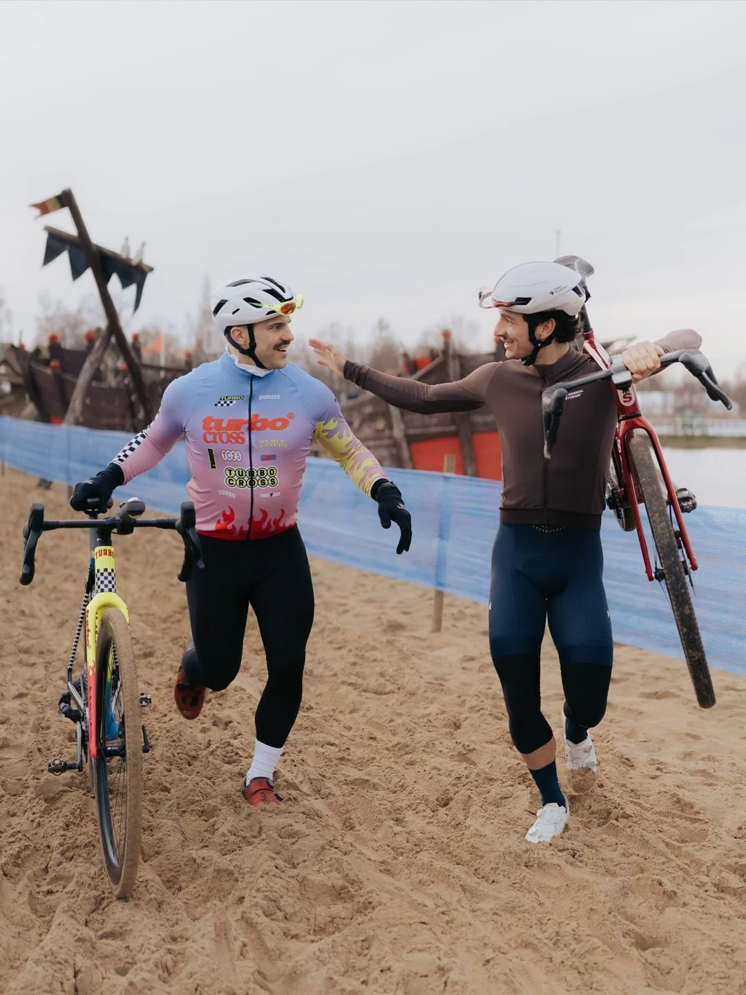 Two male cyclists in helmets share a moment of camaraderie on a sandy beach, one stepping over his bike, the other holding his bike and reaching out.