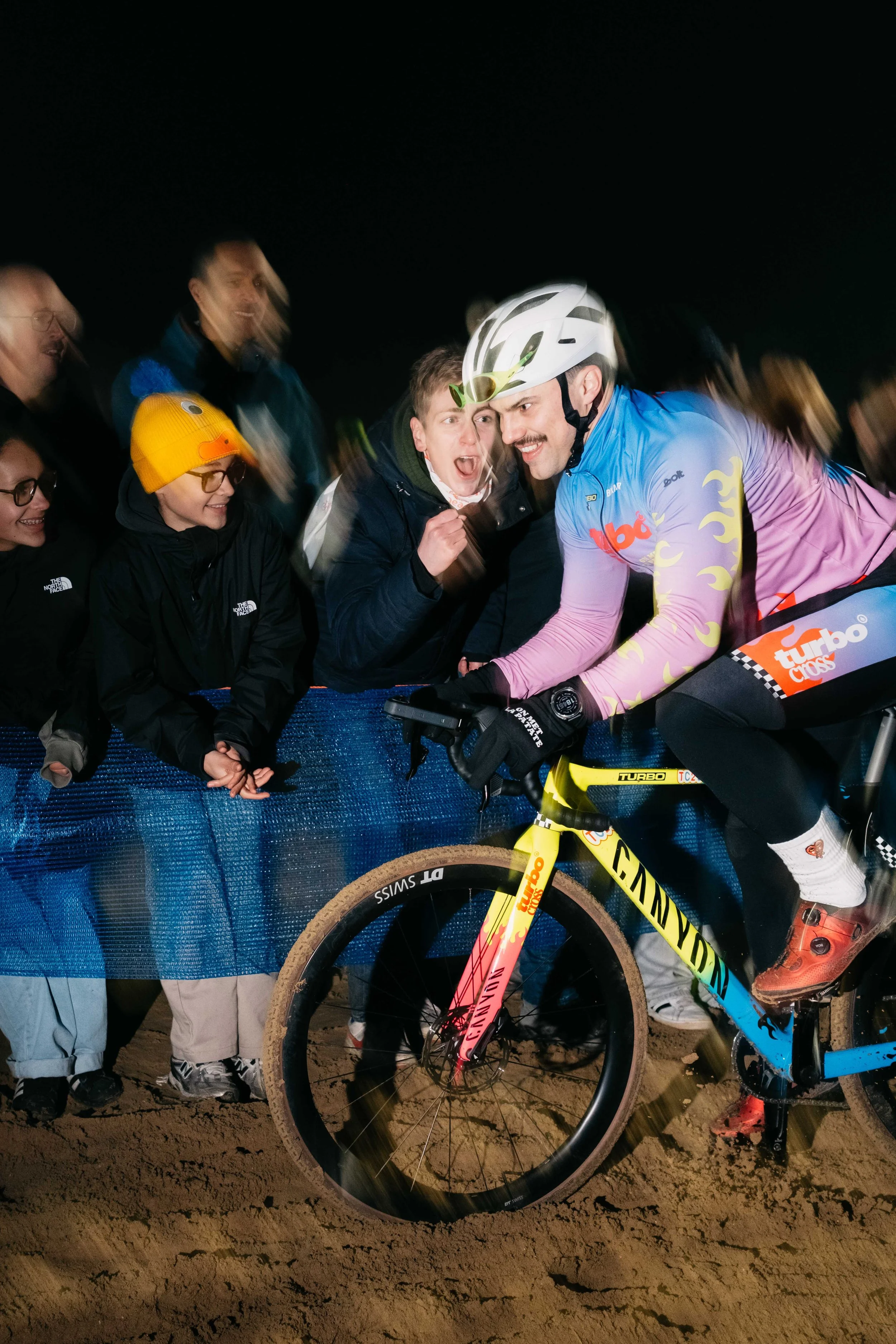 Cyclist in colorful jersey and helmet leaning on bike at night, surrounded by cheering spectators.