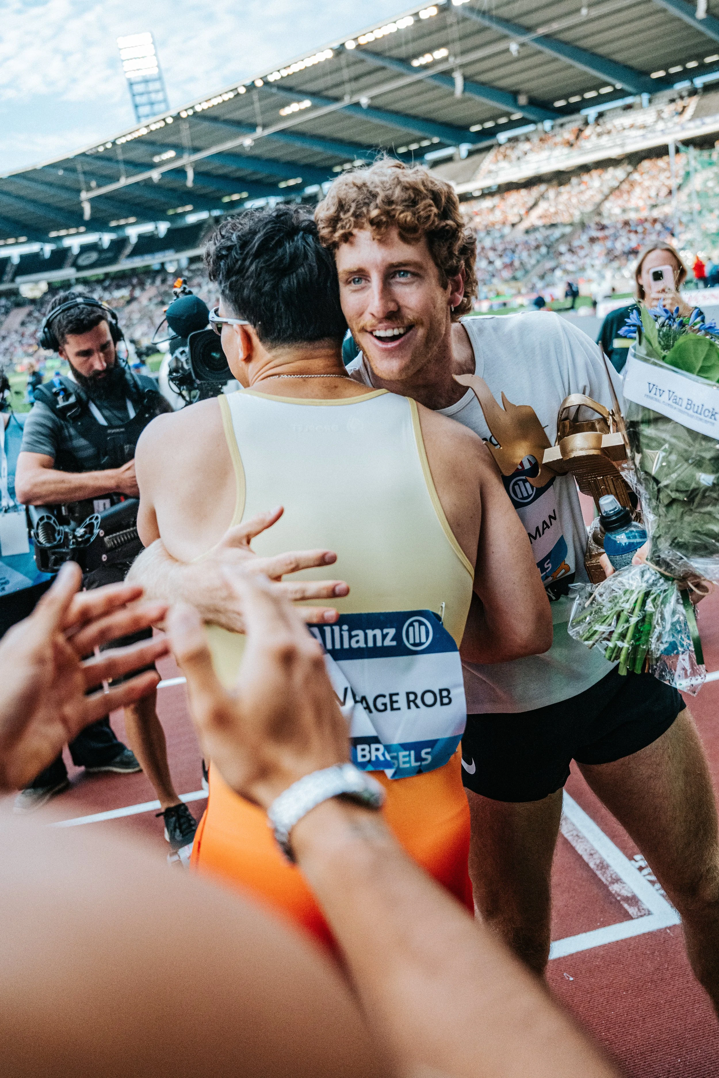 Athlete hugging another athlete on a track field, with a camera operator and spectators in the background.