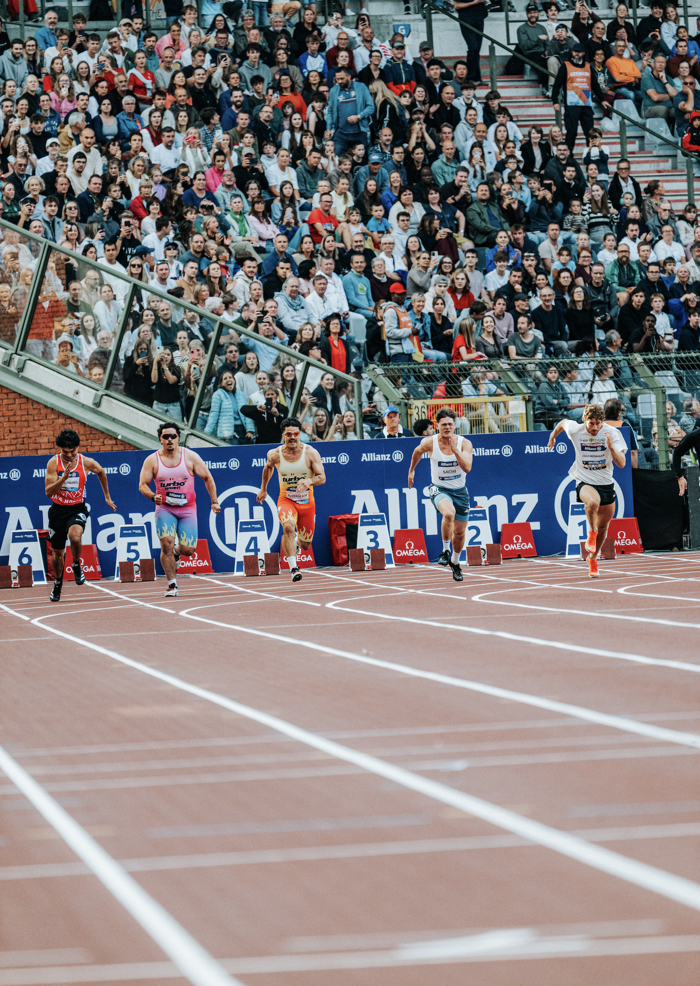 Track athletes sprinting at the start of a race with a crowd of spectators watching in the background.