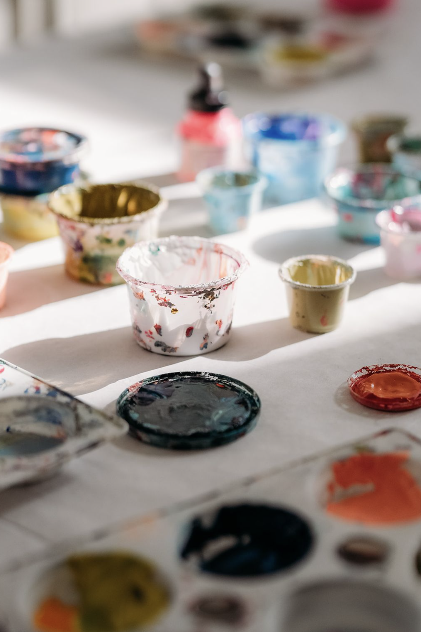 Blurred image of various open paint containers and brushes on white paper as part of a creative workshop.