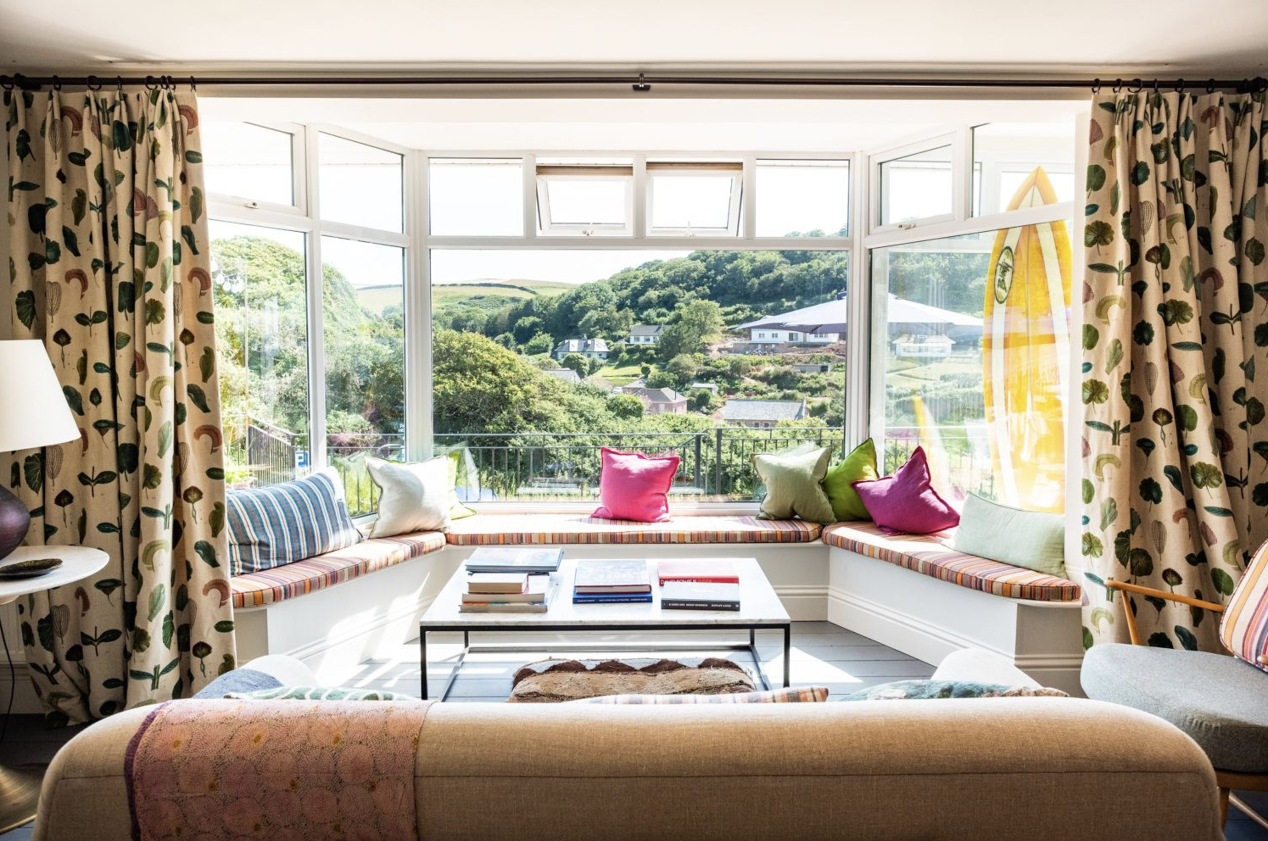 Living room with large window seating area decorated with colorful pillows, overlooking green hills and houses outside, with curtains featuring a leaf pattern.