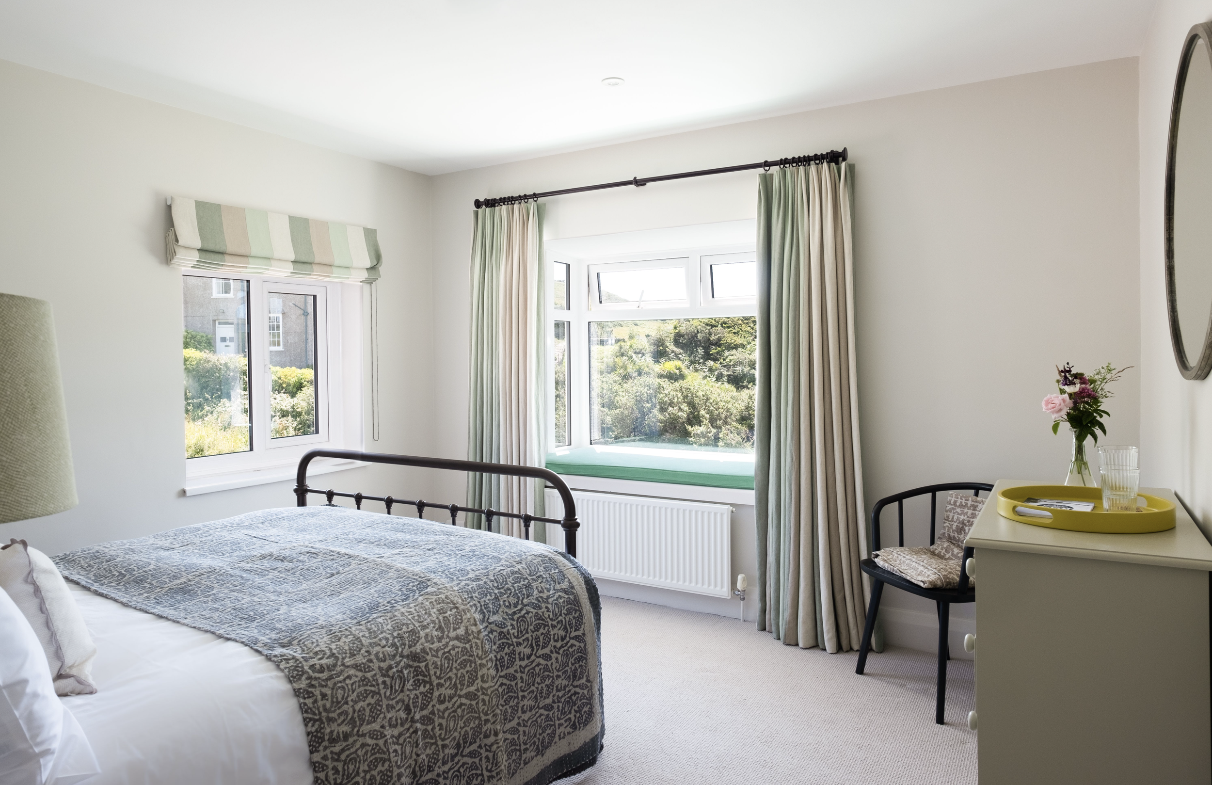 A bright bedroom with two windows, one with green and pink striped roman shade and the other with green and beige curtains, overlooking greenery outside. There's a bed with a patterned gray duvet, a bedside table with a vase of pink and purple flowers, and a yellow tray. A round mirror is mounted on the wall.