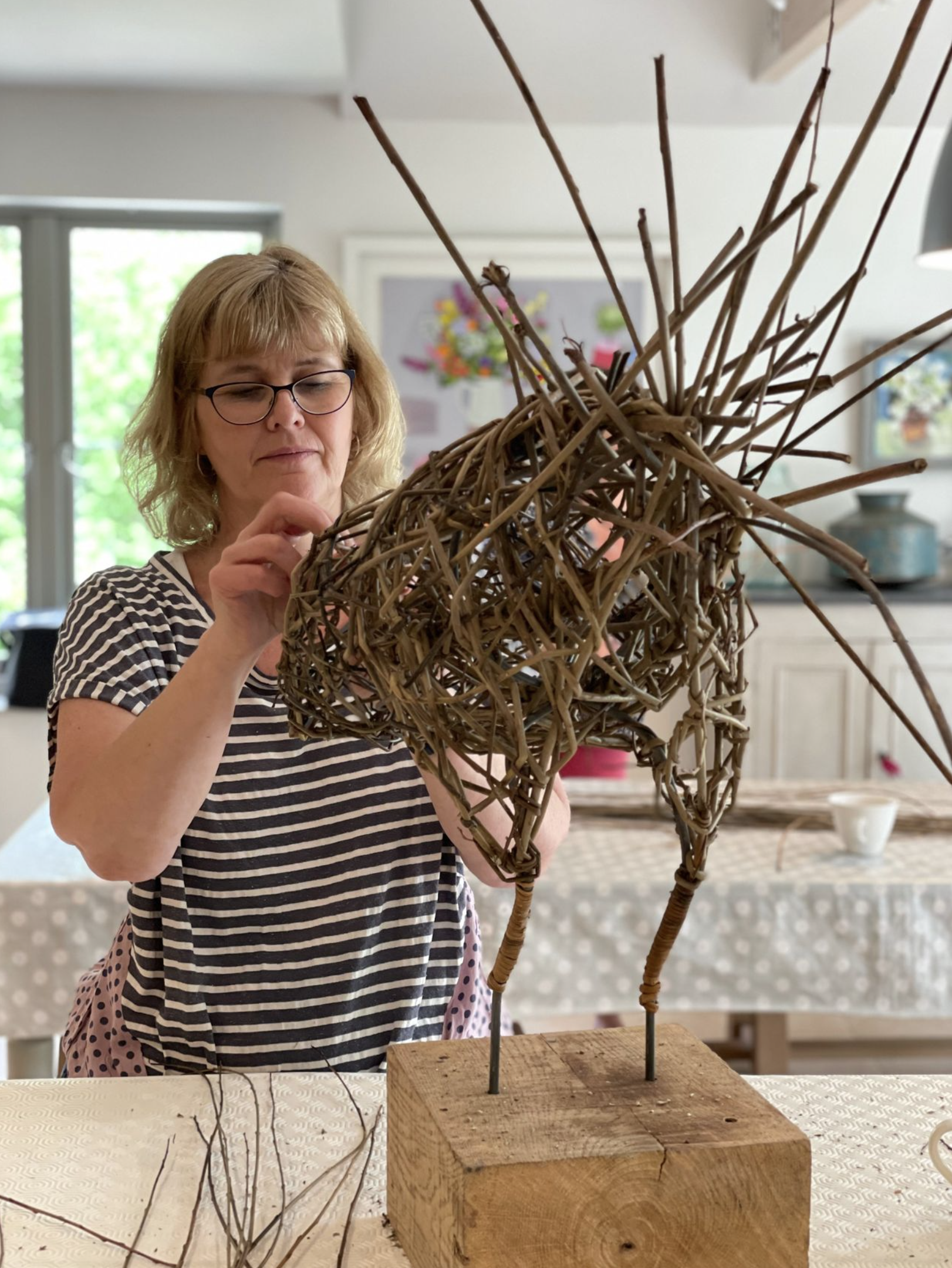 A woman with blonde hair and glasses is creating a woven chicken sculpture from willow on a wooden block in a bright space with colourful artwork in the background.