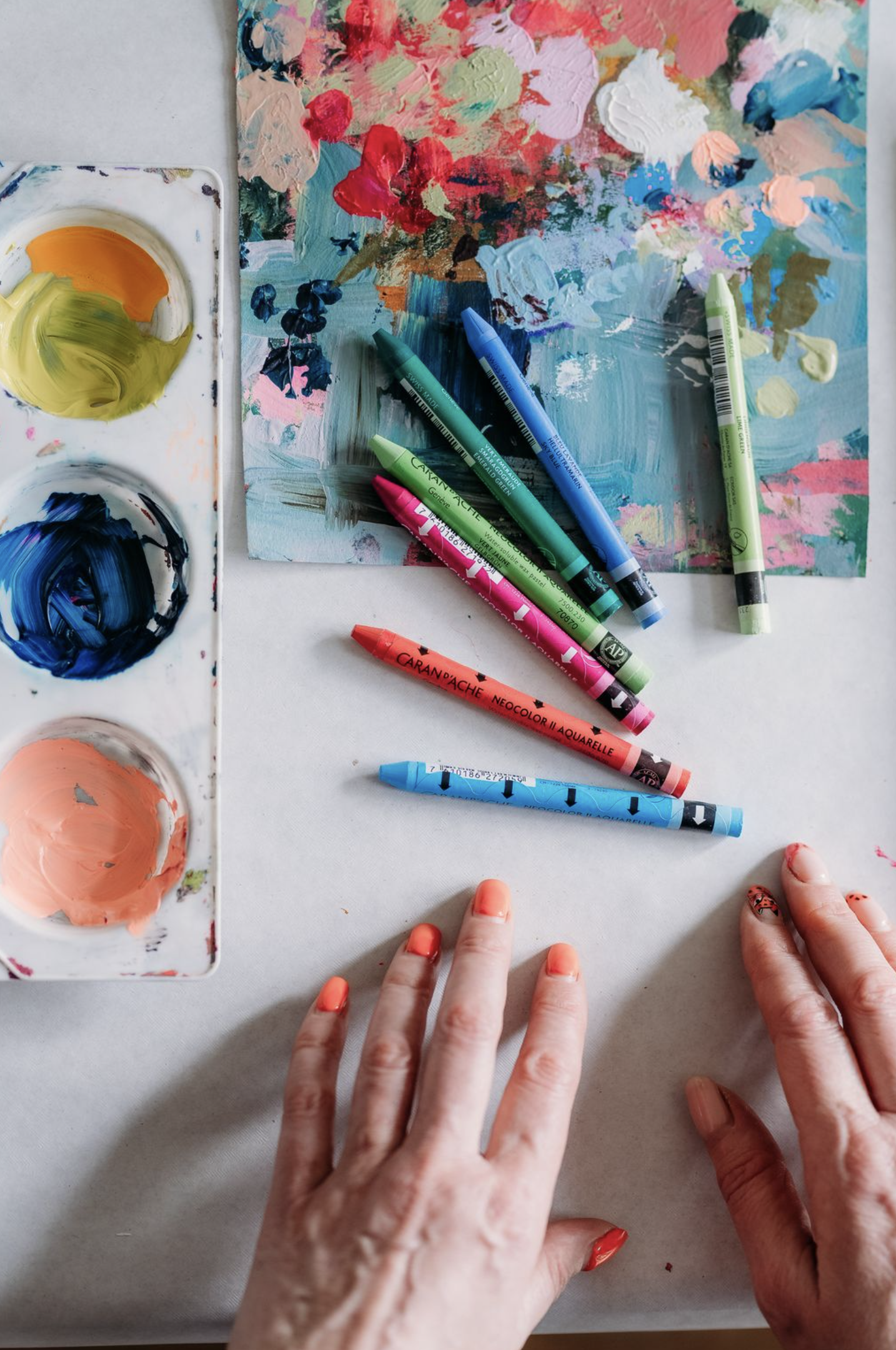Close-up of two hands with painted fingernails, resting on a white surface near a colorful abstract painting and several markers and paint palettes with various colours as part of a creative workshop.