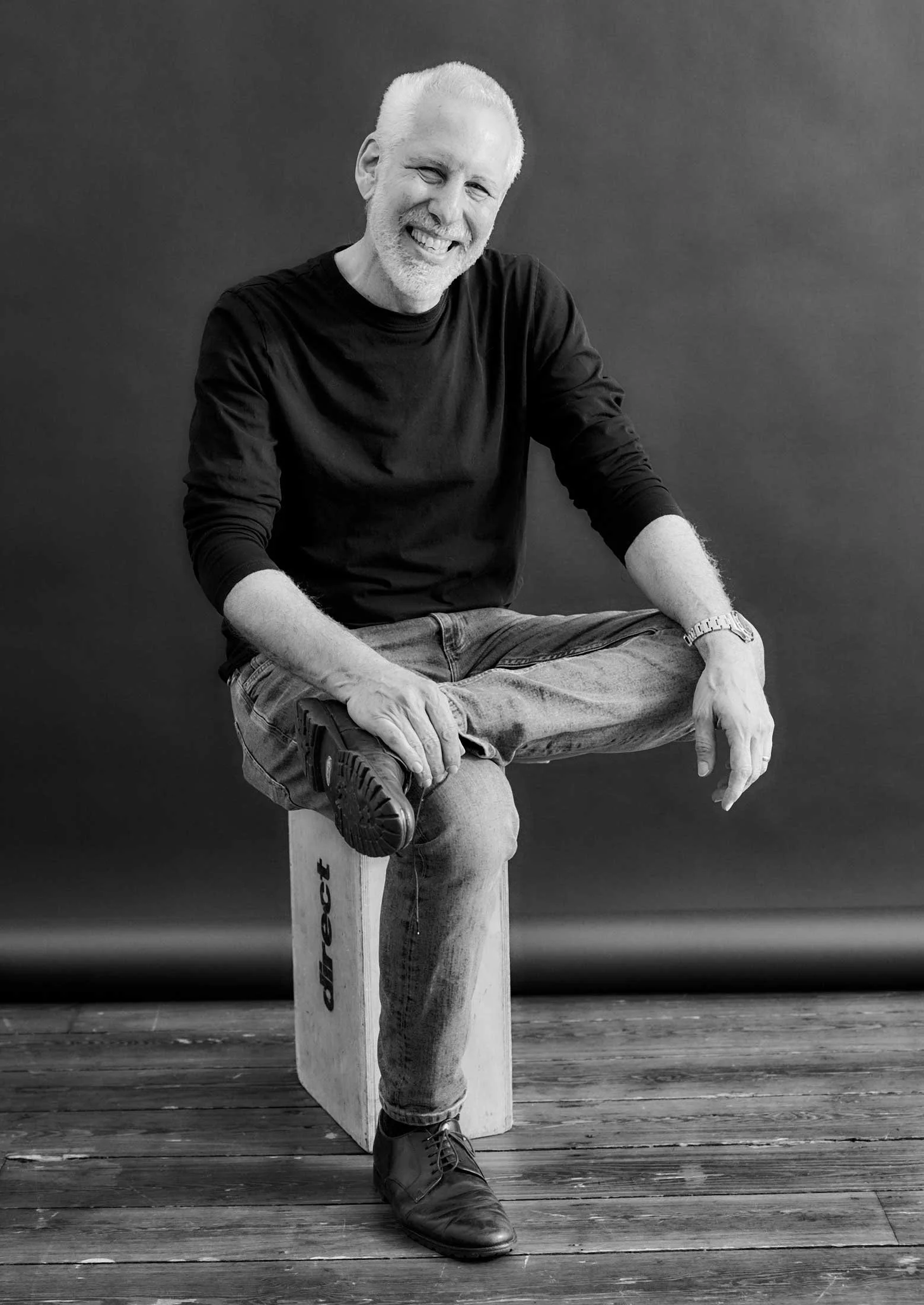 Joseph Montezinos wearing a black shirt and jeans, sitting on a wooden stool with one leg crossed over the other, against a plain dark background.