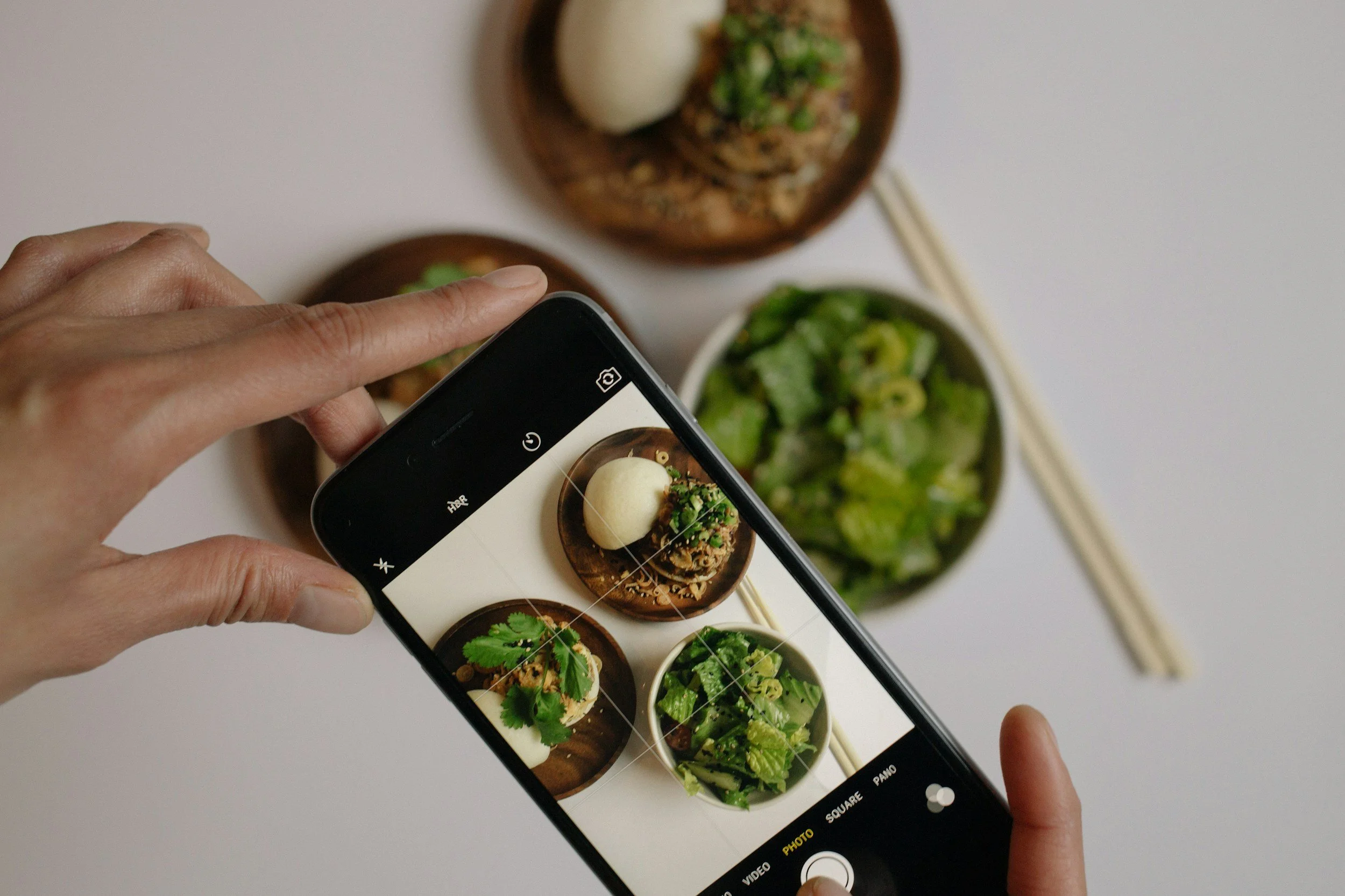 Person taking a photo of small bowls of Thai food in a Hotel in Bangkok using a smartphone, with chopsticks beside the bowls on a white surface.