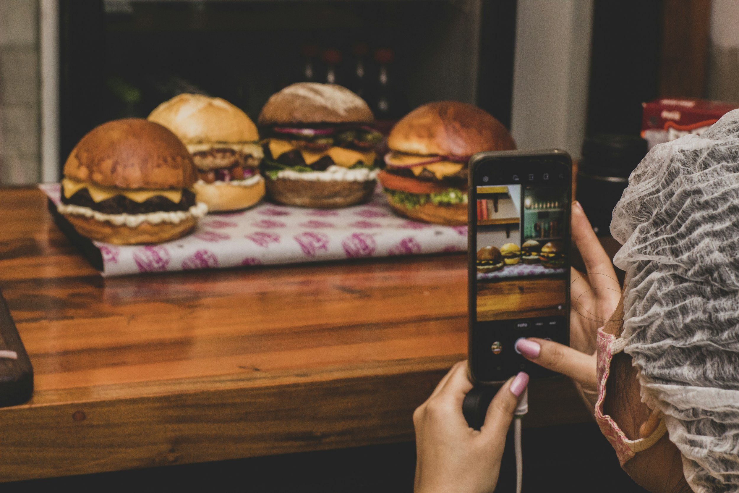 Person taking a photo of four burgers placed on a pink and white paper on a wooden table, with the person's hand and a smartphone visible in the foreground.