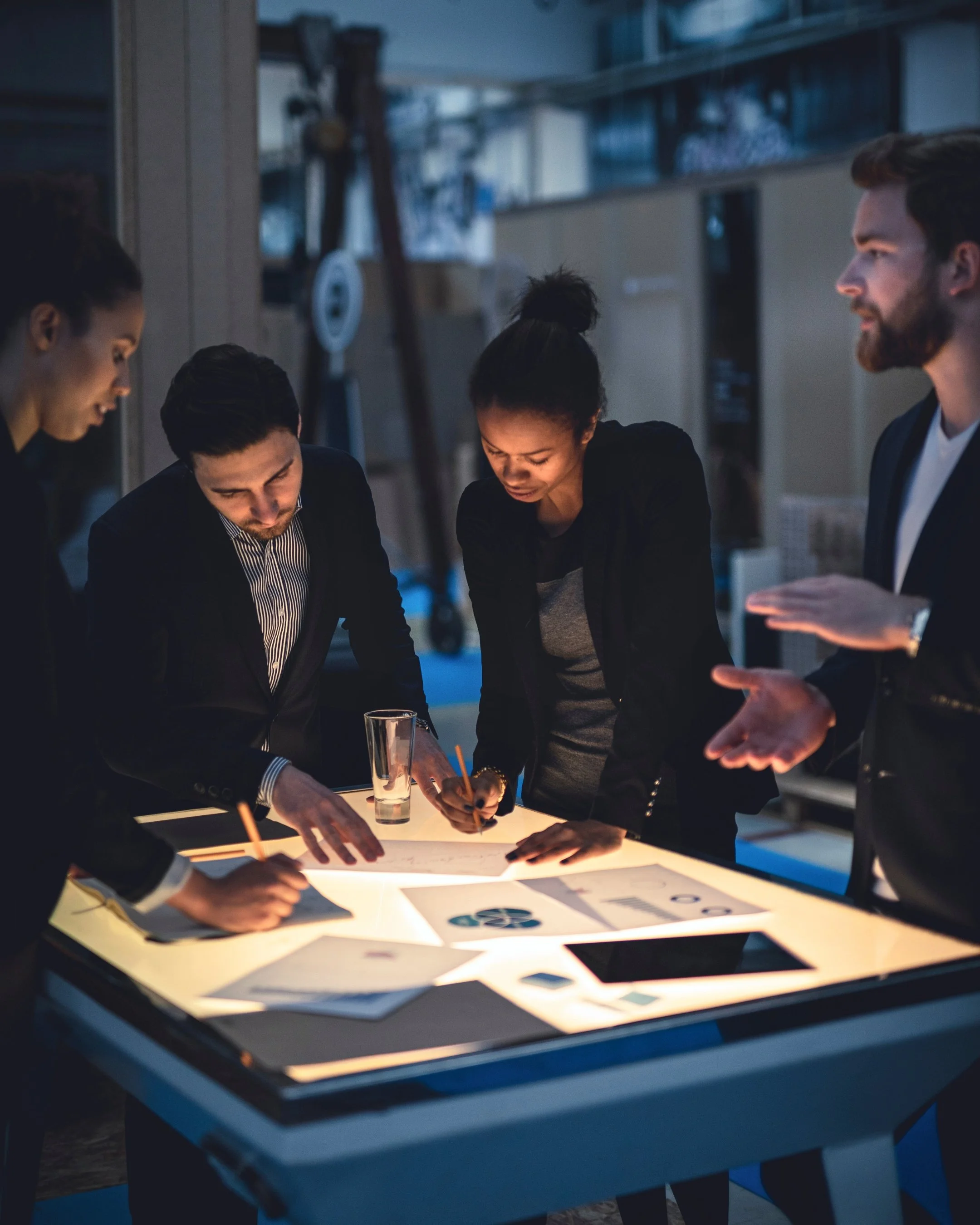 Group of four people gathered around a light table with various charts and graphs, discussing and working on a project in a modern indoor setting.