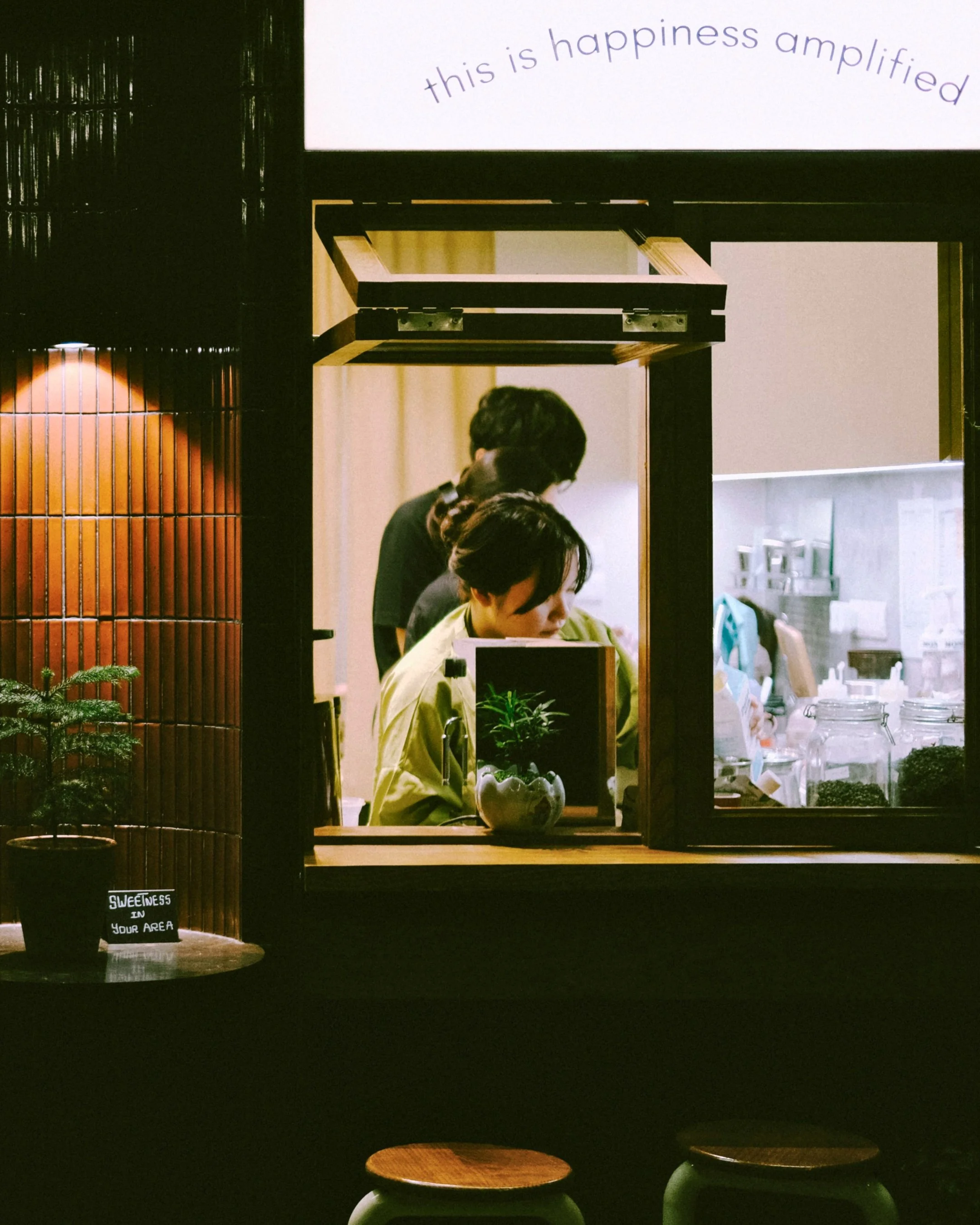 A view of a small indoor café or juice bar through a window with wooden frames. Inside, there are two women preparing drinks or food at a counter, with shelves holding jars and bottles. A sign above reads 'this is happiness amplified.' Outside, a small round wooden table with two stools is visible, one with a small potted plant on it.