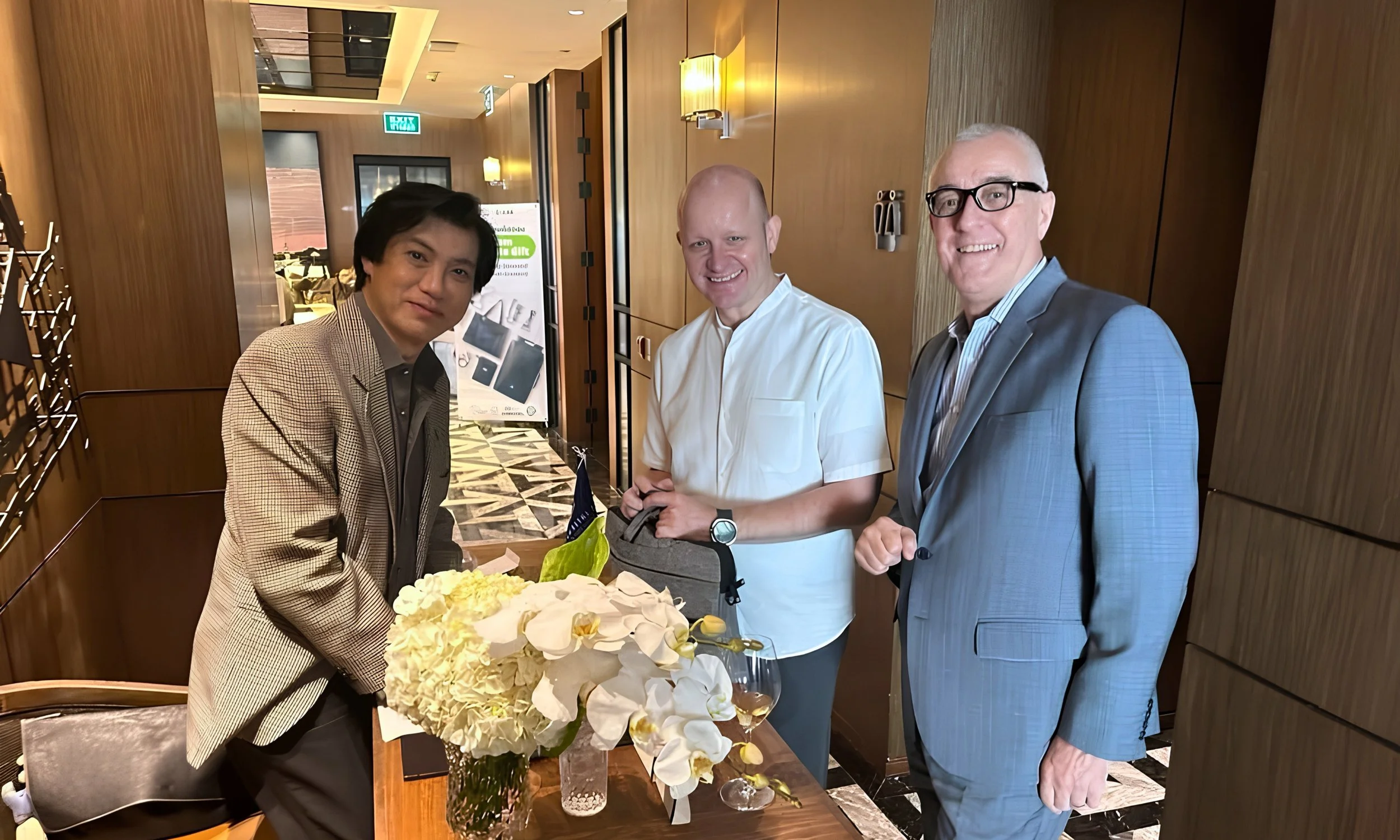 Three men standing behind a table with a bouquet of flowers in a restaurant or hotel lobby, smiling at the camera.