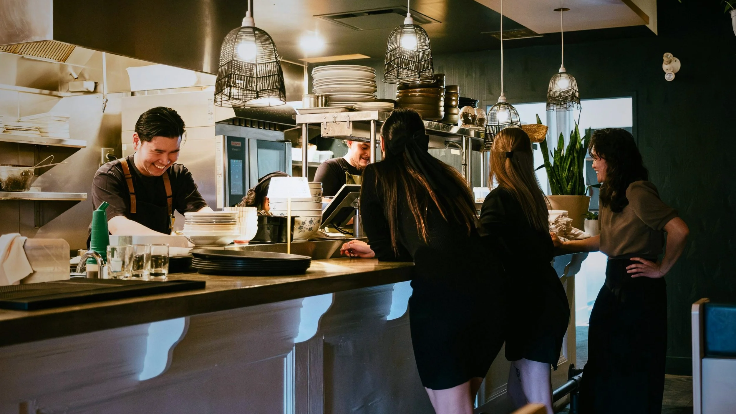 The image shows a busy restaurant kitchen with male and female staff preparing food and interacting at the counter. The kitchen is modern with hanging pendant lights, shelves with plates, and various kitchen equipment.