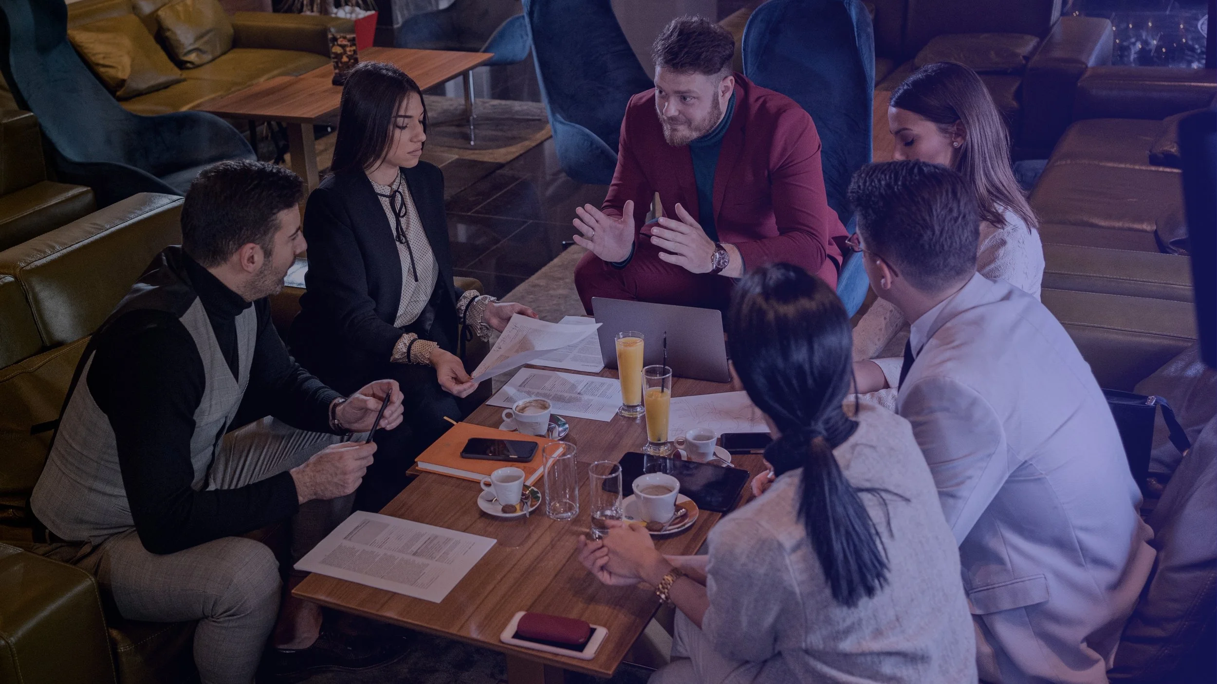 A group of seven diverse people, including men and women, sitting around a wooden table in a cozy, dimly lit cafe or restaurant, engaged in a meeting or discussion, with papers, notebooks, and drinks on the table.
