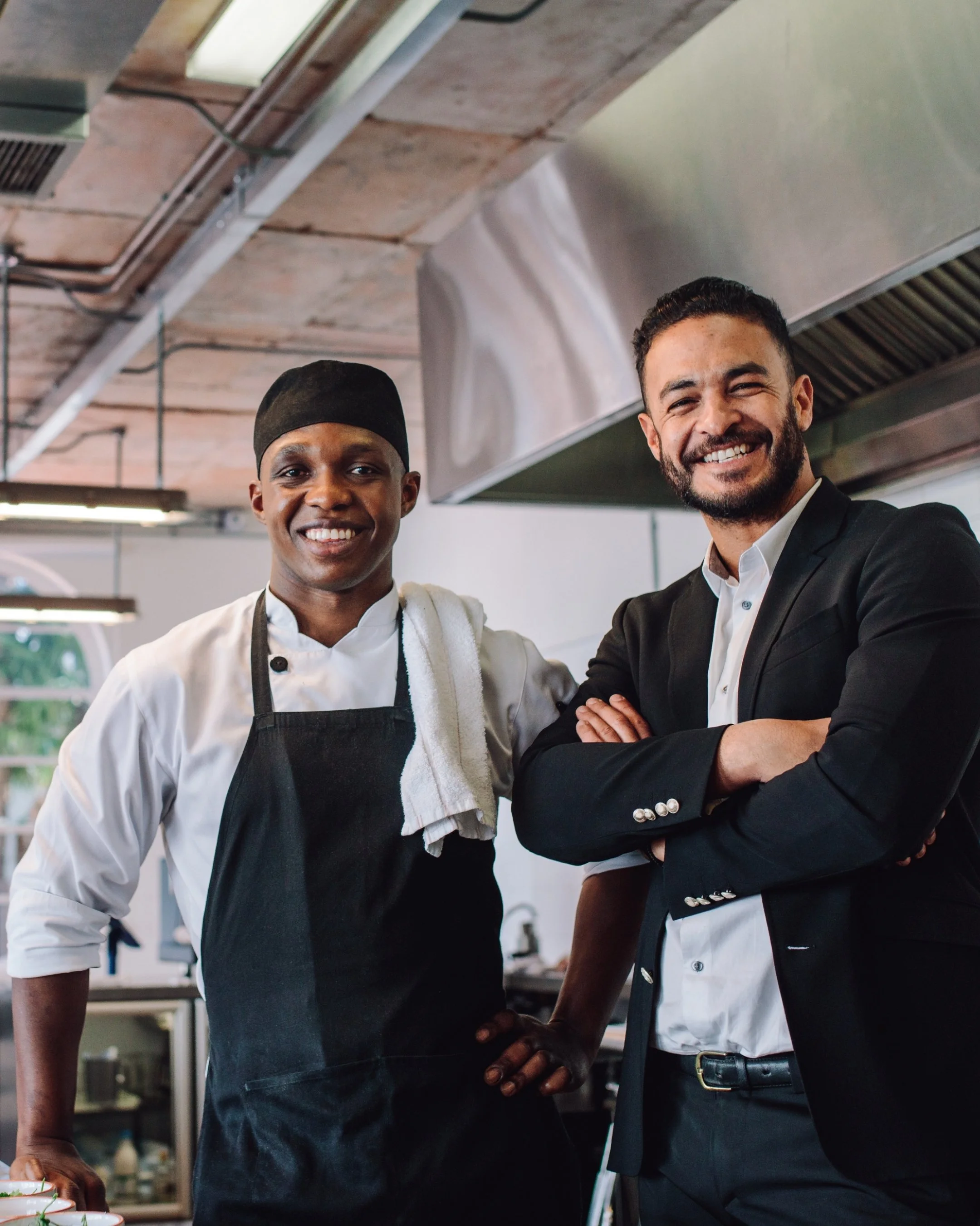Two men smiling and standing close together in a restaurant or kitchen setting. The man on the left is wearing a chef's uniform with a black apron and has a towel draped over his shoulder. The man on the right is dressed in a dark suit with a white shirt, arms crossed, and both men are looking at the camera.