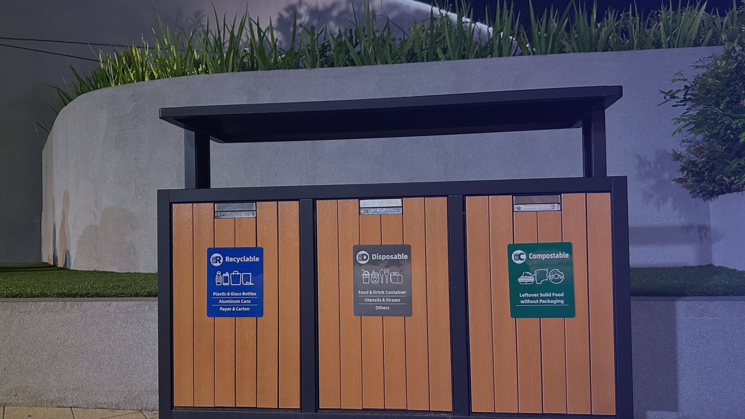 An outdoor recycling station with three labeled sections for recyclable, disposable, and compostable items, against a background of a curved concrete wall and greenery.
