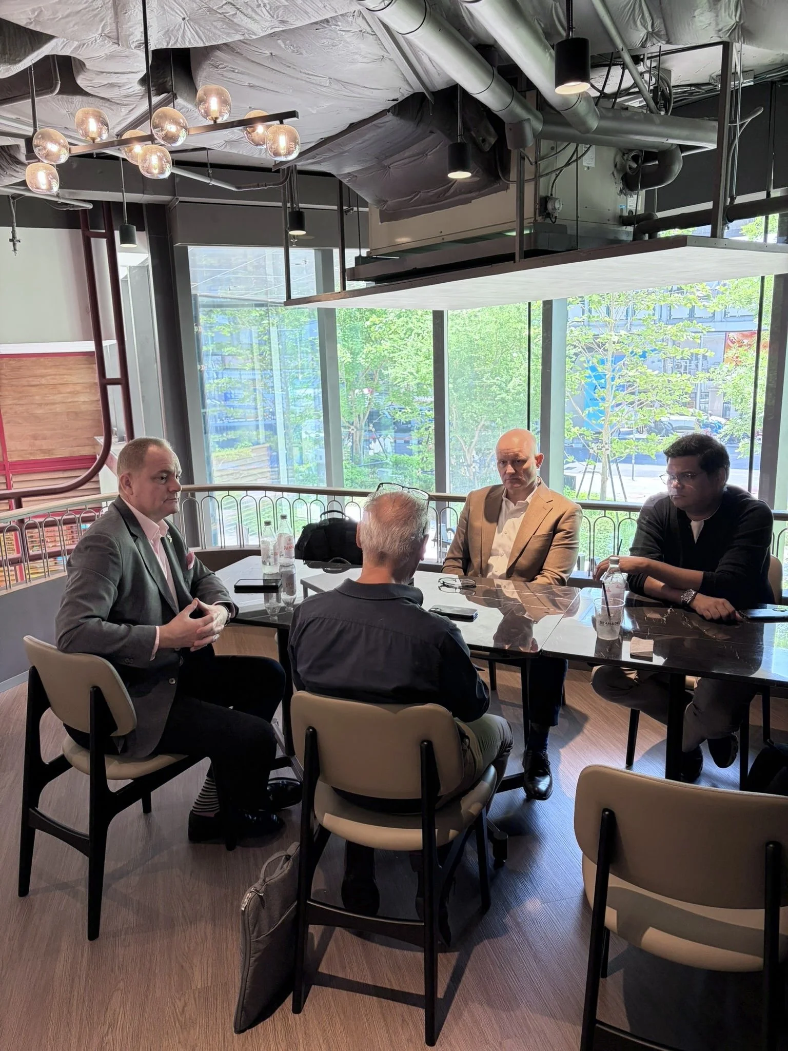 A group of five hospitality experts sitting around a rectangular table in a modern restaurant or cafe with large windows in the background, some with bottles and drinks on the table, engaged in conversation.