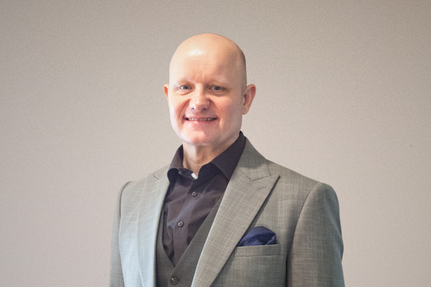 Smiling bald man in a white shirt standing indoors with greenery and city buildings in the background.