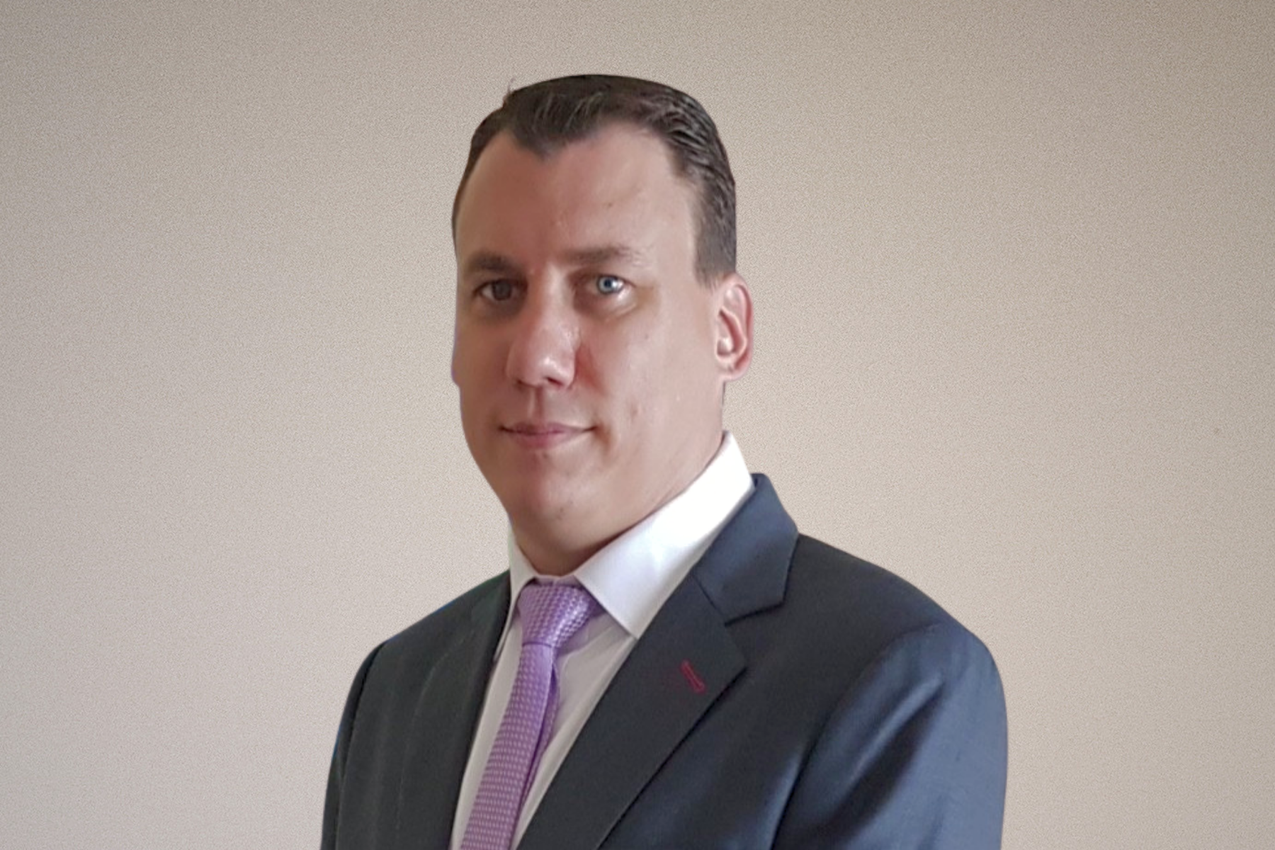 A man with slicked-back dark hair, wearing a dark suit, white shirt, and light purple tie, posing indoors in front of a framed picture of a roller coaster at an amusement park.