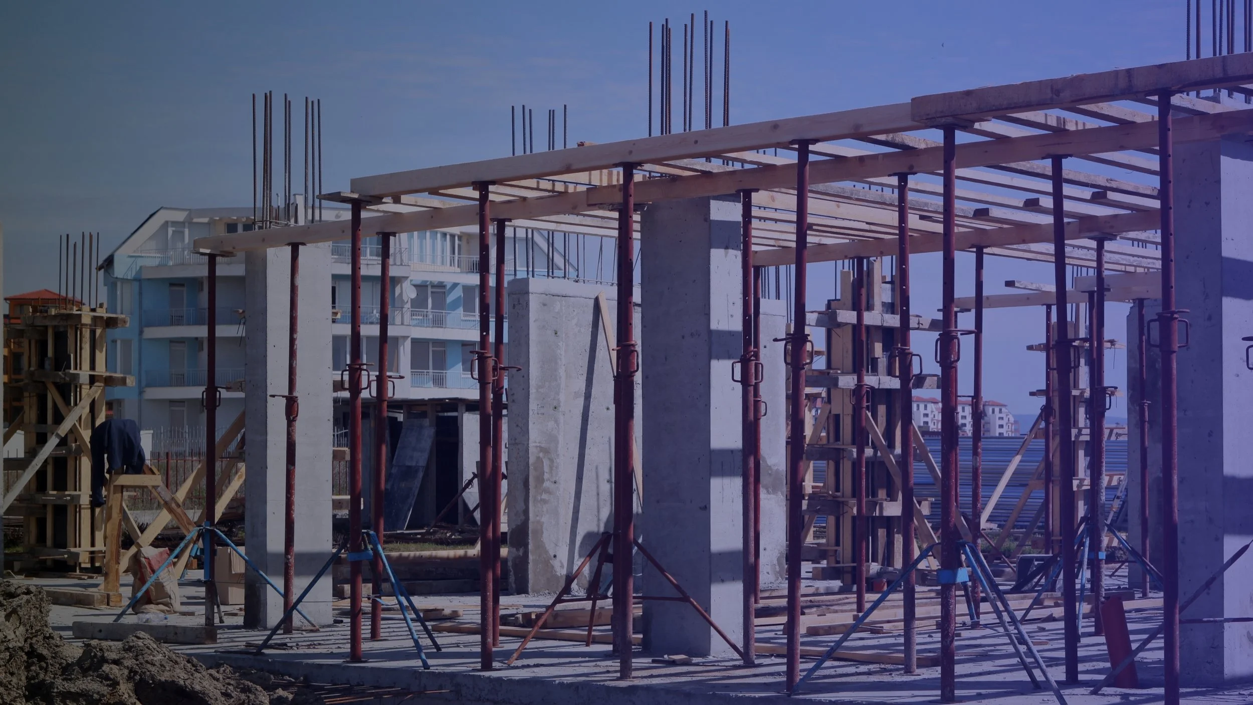 Construction site with concrete columns and wooden beams supported by red scaffolding, with a modern residential building in the background under a blue sky.