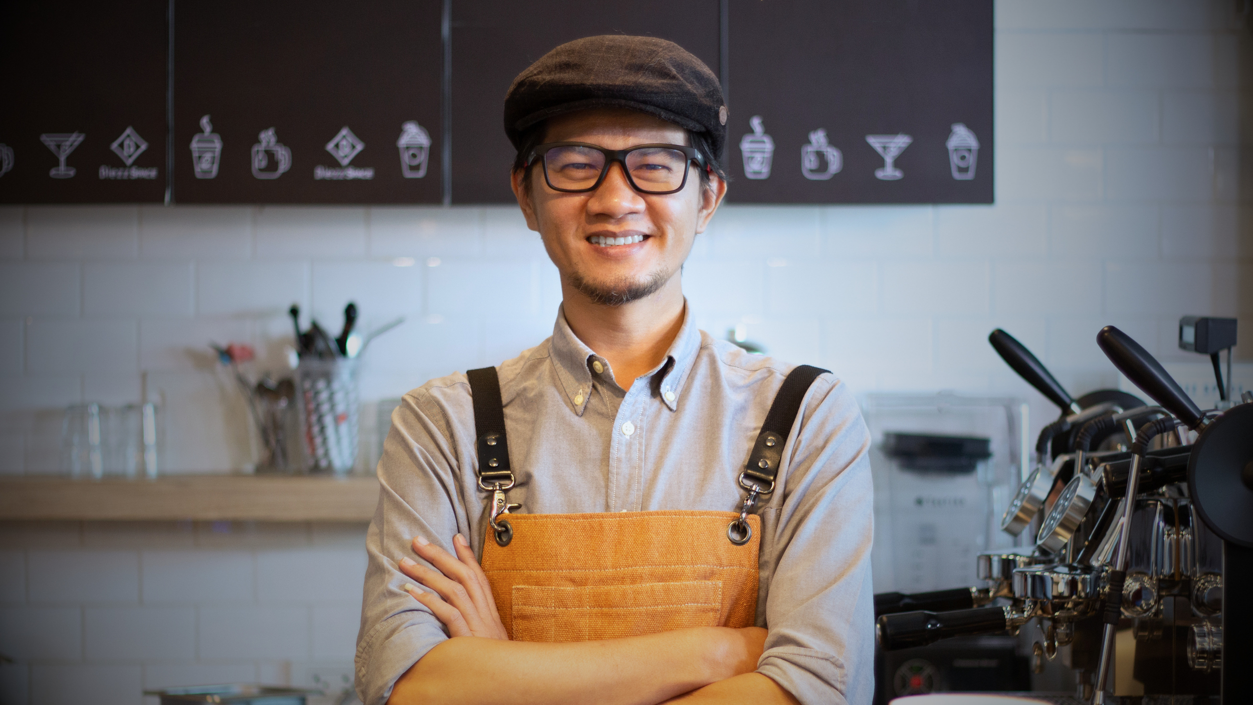 A smiling barista with glasses, a flat cap, and an orange apron standing with arms crossed in a coffee shop kitchen.