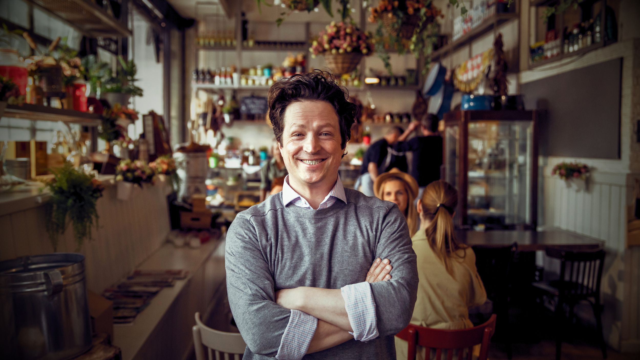 A man with dark, curly hair and a gray sweater standing with arms crossed inside a cozy café, smiling at the camera. The café has wooden shelves with jars, flowers, and decorative items, with others sitting and chatting in the background.