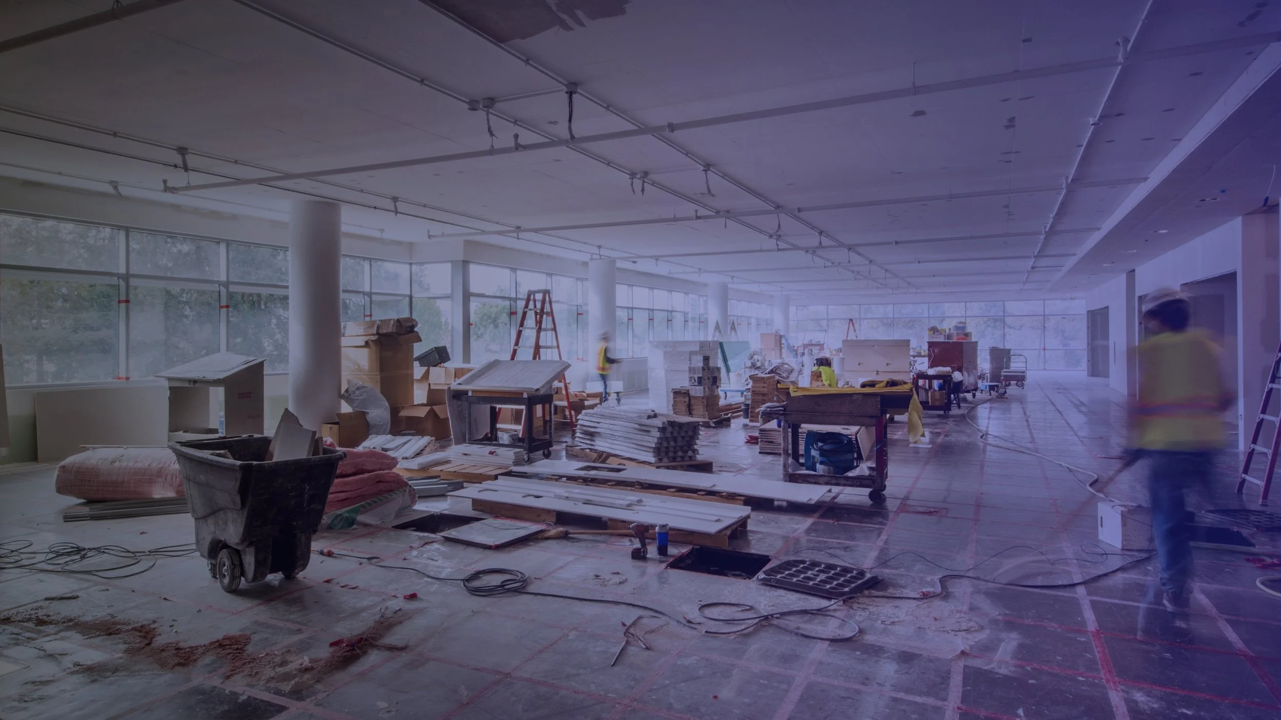 Interior of a building under construction with large windows, construction materials, ladders, and workers.