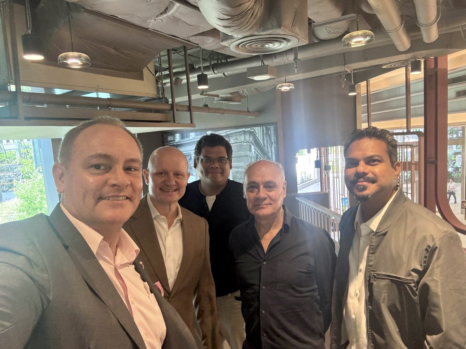 Five men are standing together in a restaurant or cafe, smiling at the camera with a city street visible through the windows in the background.