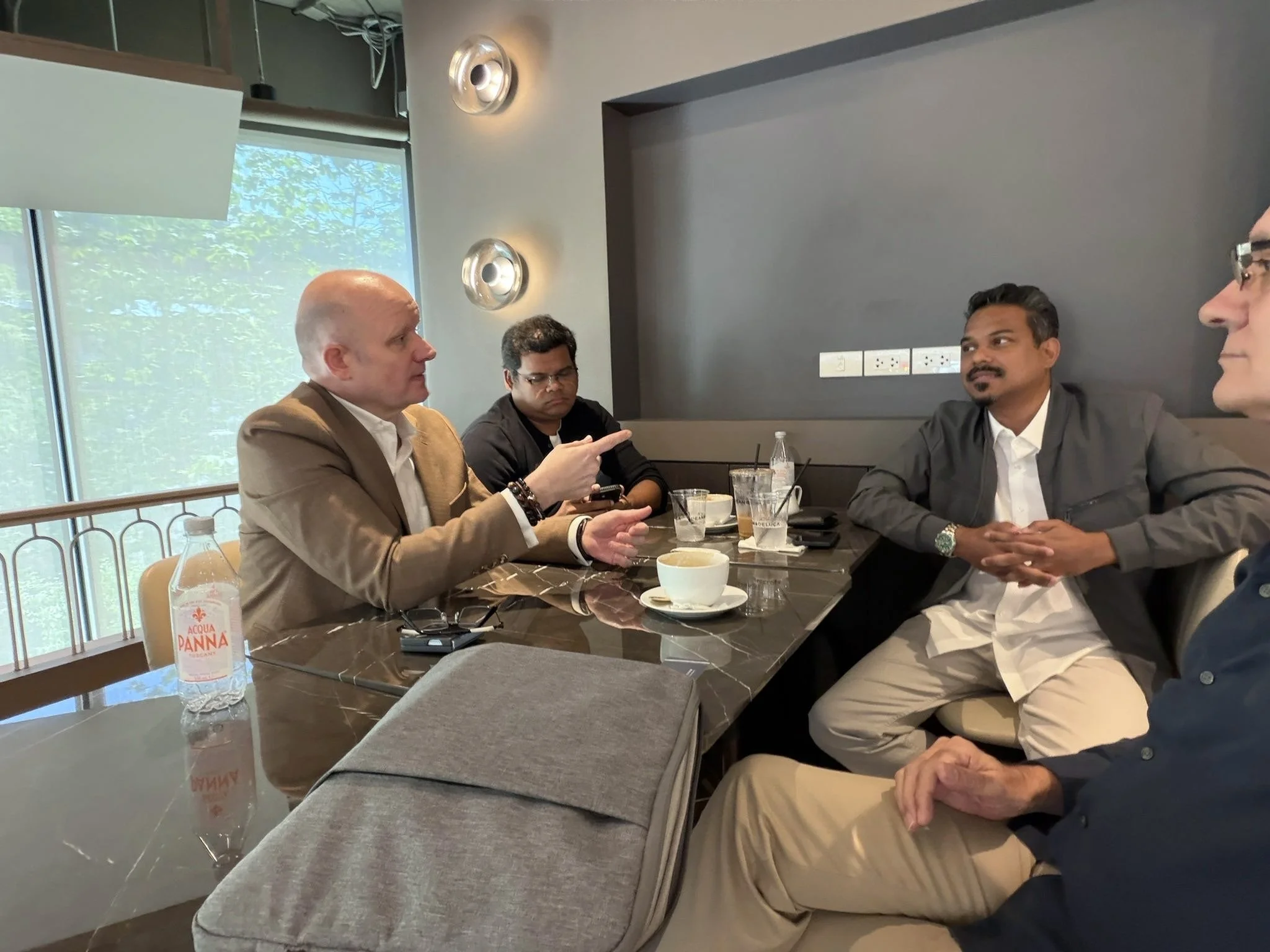 Four men sitting at a table in a restaurant having a conversation. One man is gesturing with his hand, and another man is looking at him. The table has drinks, a cup, and a bottle of water. There is a window with greenery outside and modern wall lights behind them.