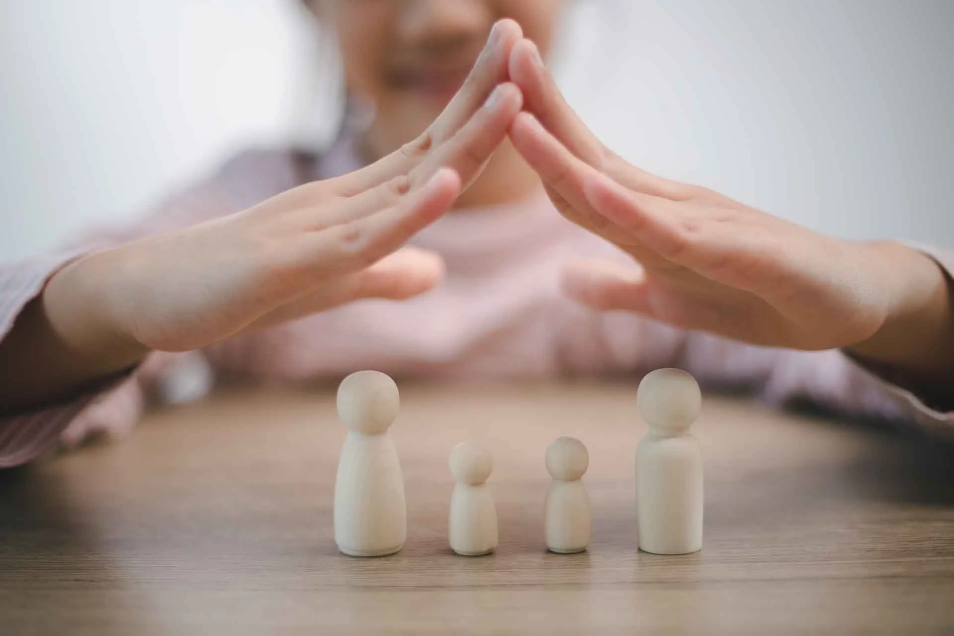 Una persona fa un gesto di protezione con le mani sopra quattro figure di legno di diverse dimensioni disposte in ordine crescente da sinistra a destra, simboleggiando protezione o supporto.