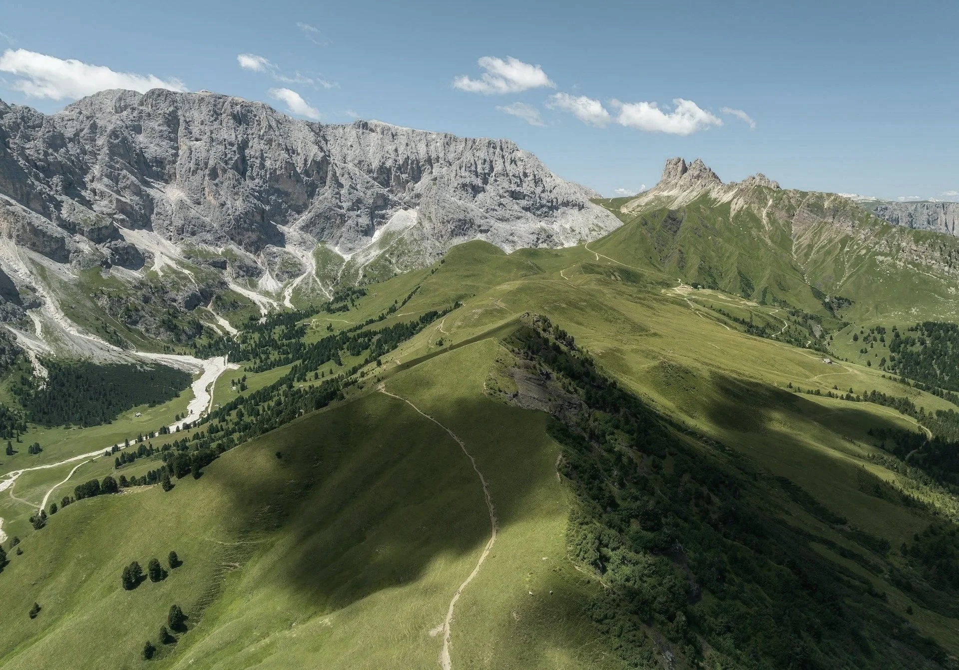 Berglandschaft mit grünen Hügeln, Wiesen und einem Tal vor hohen, kargen Bergen unter einem blauen Himmel mit Wolken.
