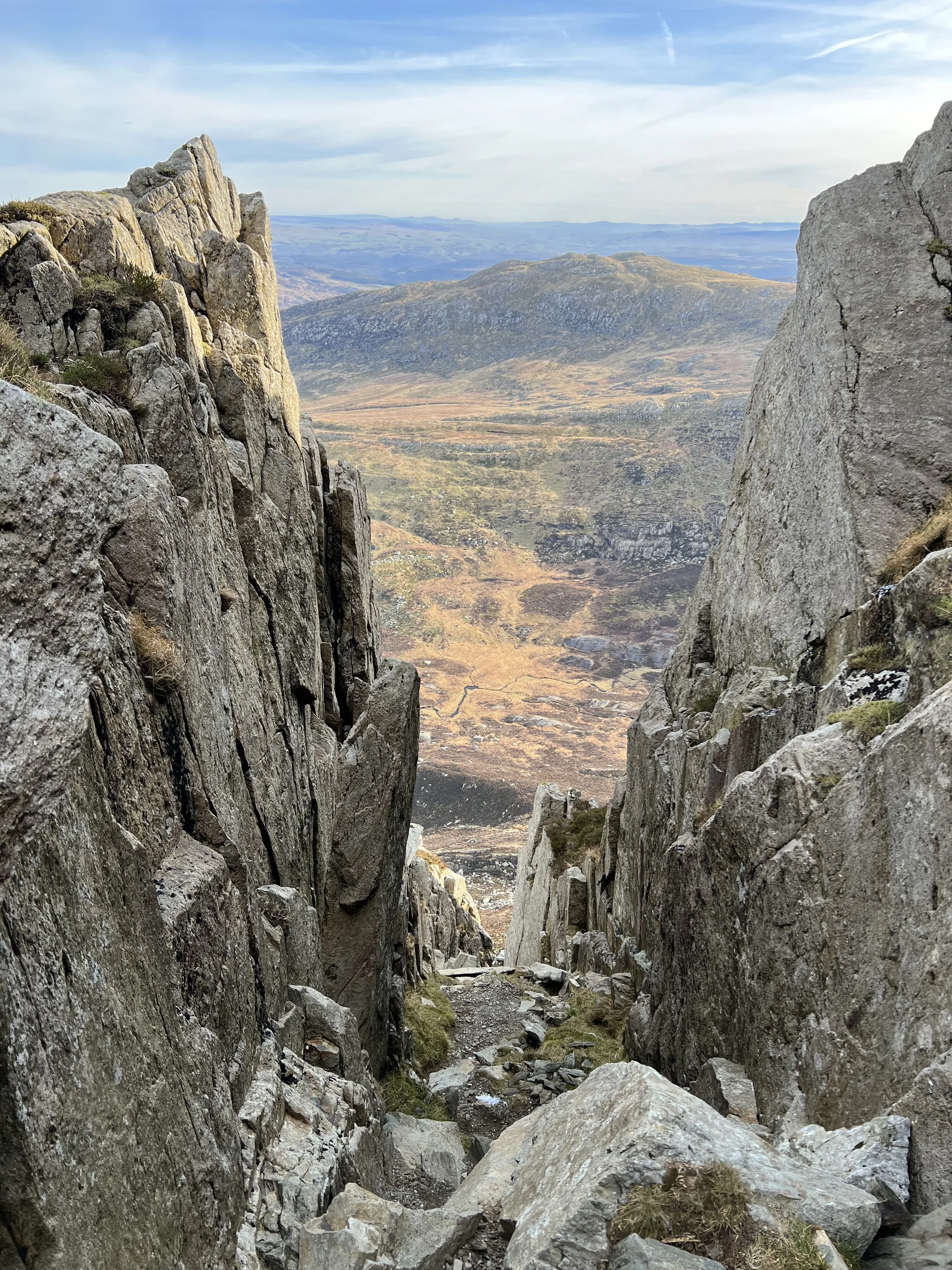 A steep rocky mountain trail between two large rock formations overlooking a valley with distant hills under a blue sky.